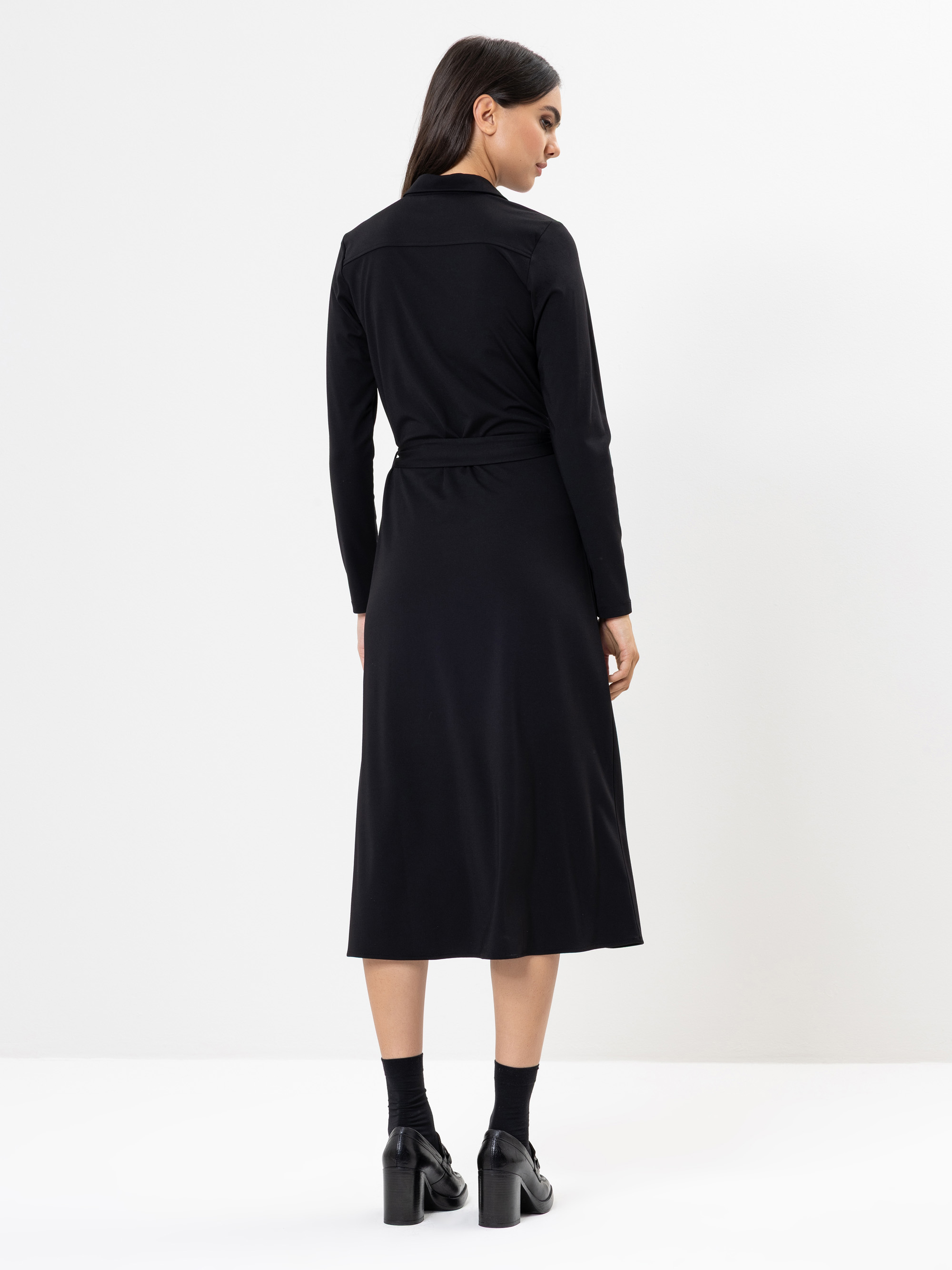 Frau, die mit dem Rücken zur Kamera steht und ein langärmeliges, schwarzes, mittellanges Kleid, schwarze Socken und schwarze Schuhe mit Absätzen trägt, vor einem schlichten weißen Hintergrund.