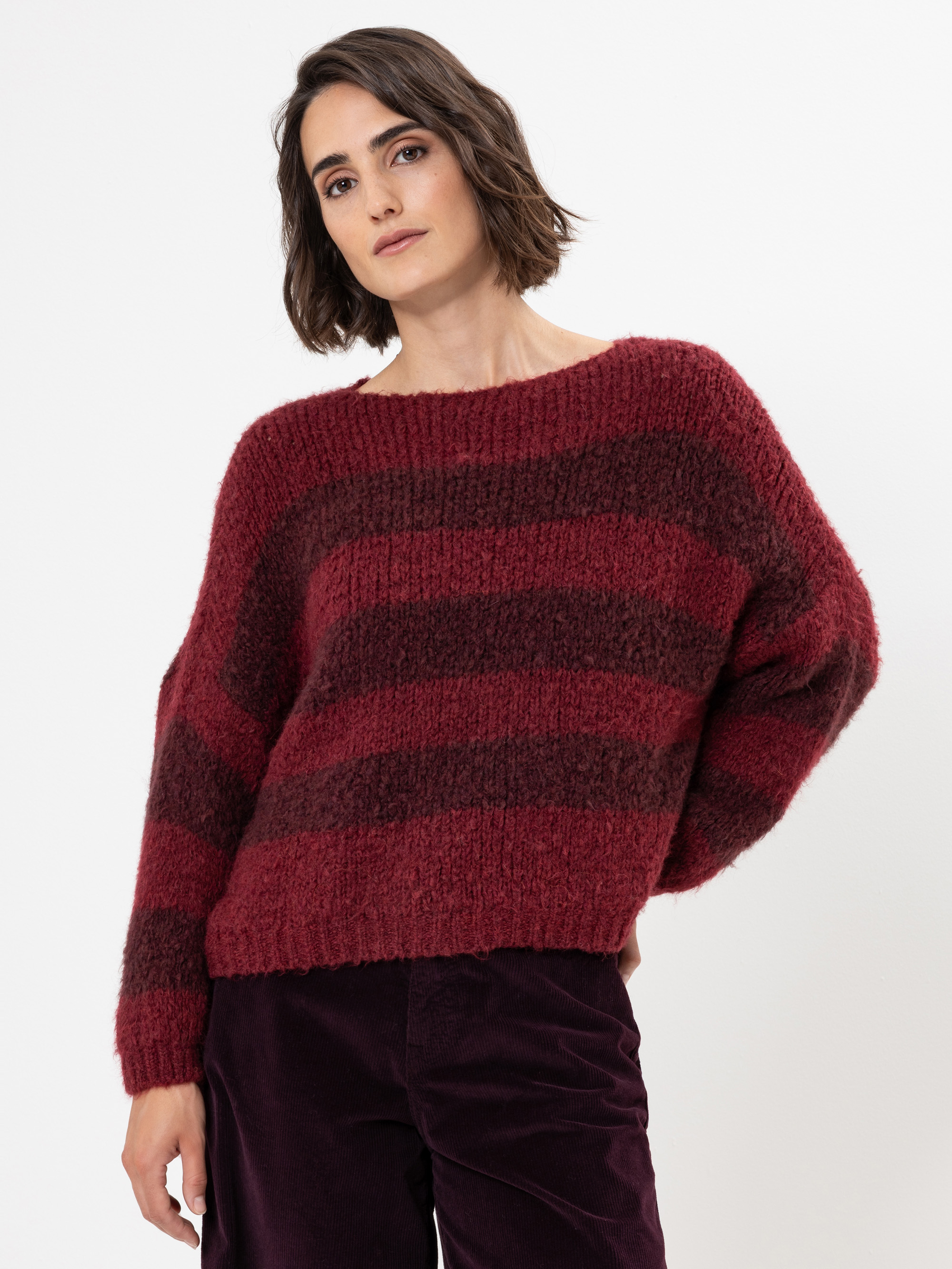 Eine Person mit kurzen braunen Haaren, die einen lockeren, kastanienbraunen und dunkelrot gestreiften Pullover und eine dunkle Hose trägt, steht vor einem schlichten weißen Hintergrund und blickt mit entspanntem Blick in die Kamera.