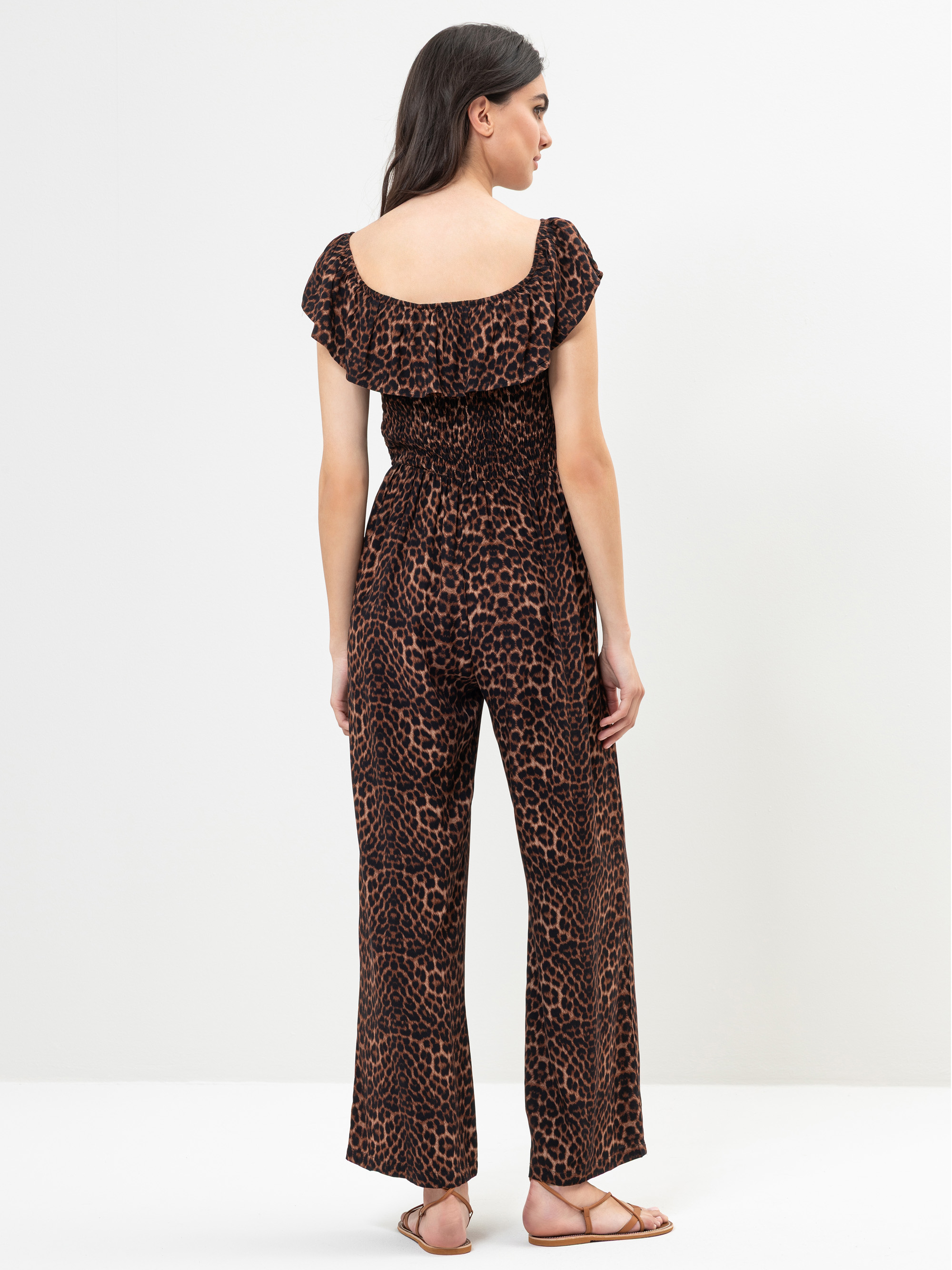 Eine Frau steht mit dem Gesicht zur Wand und trägt einen braunen und schwarzen schulterfreien Overall mit Leopardenmuster, kurzen Ärmeln und weiten Hosenbeinen. Sie hat langes dunkles Haar und trägt flache Sandalen. Der Hintergrund ist einfarbig und we