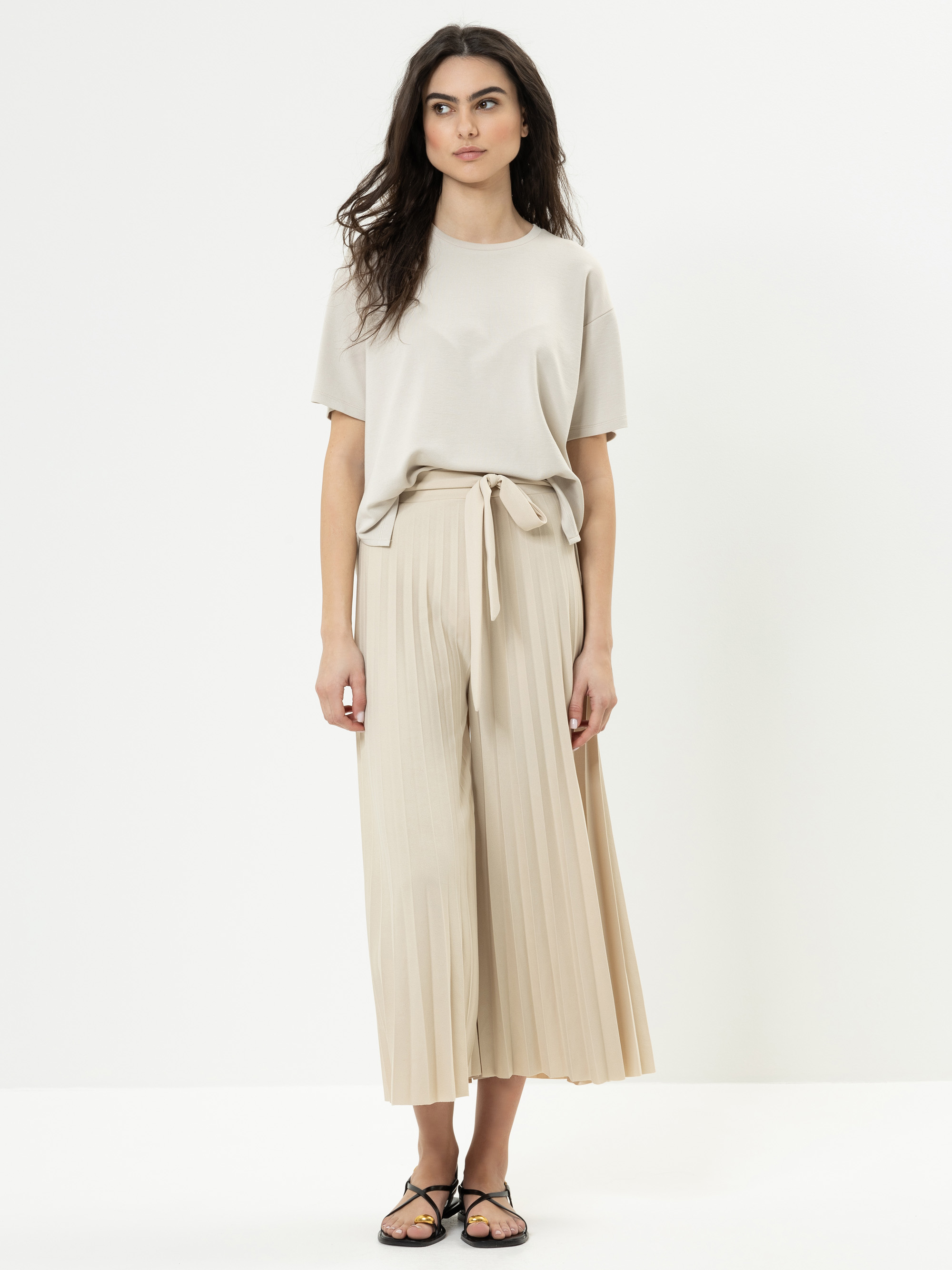 Eine Frau steht vor einem schlichten weißen Hintergrund und trägt ein beigefarbenes kurzärmeliges T-Shirt, eine hellbeige Plissee-Culottes mit Bindegürtel und schwarze Sandalen. Sie hat langes, dunkles Haar und einen neutralen Gesichtsausdruck.