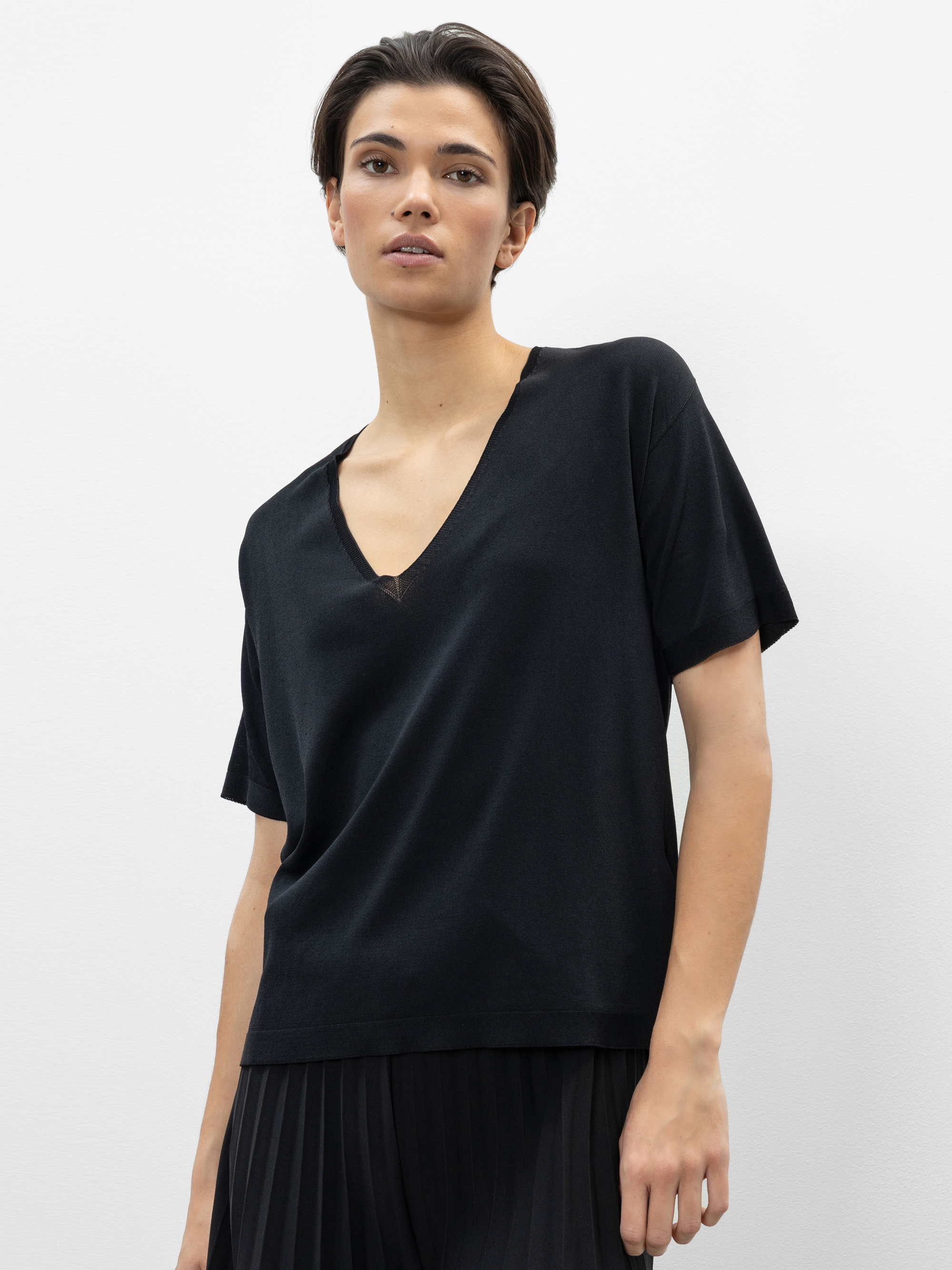 Eine Person mit kurzen dunklen Haaren trägt ein locker sitzendes schwarzes Oberteil mit V-Ausschnitt und eine schwarze Bundfaltenhose und steht vor einem schlichten weißen Hintergrund.