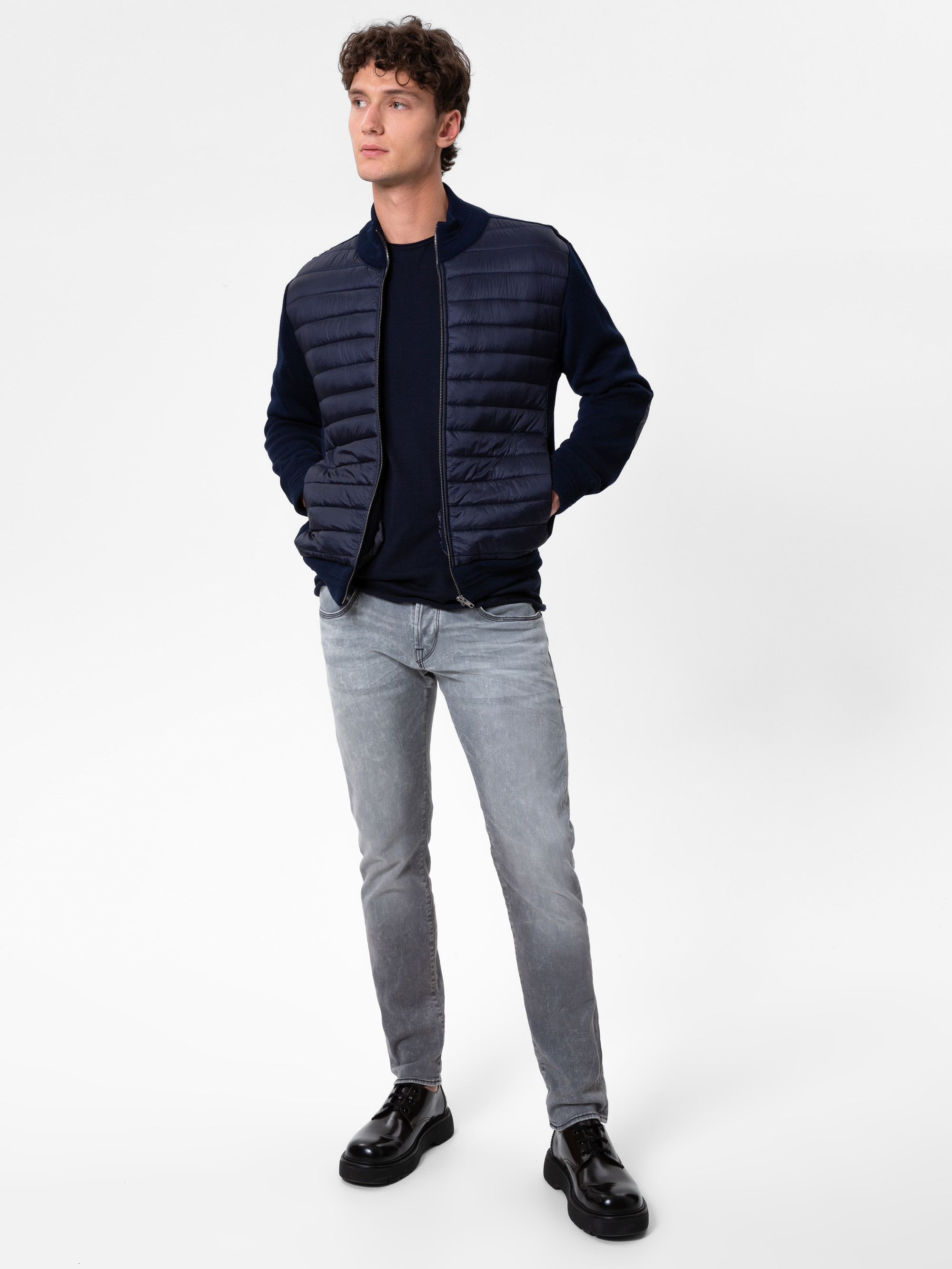 Ein Mann, der eine marineblaue wattierte Jacke, einen dunklen Pullover, hellgraue Jeans und schwarze Schuhe trägt, steht mit den Händen in den Taschen vor einem schlichten weißen Hintergrund.