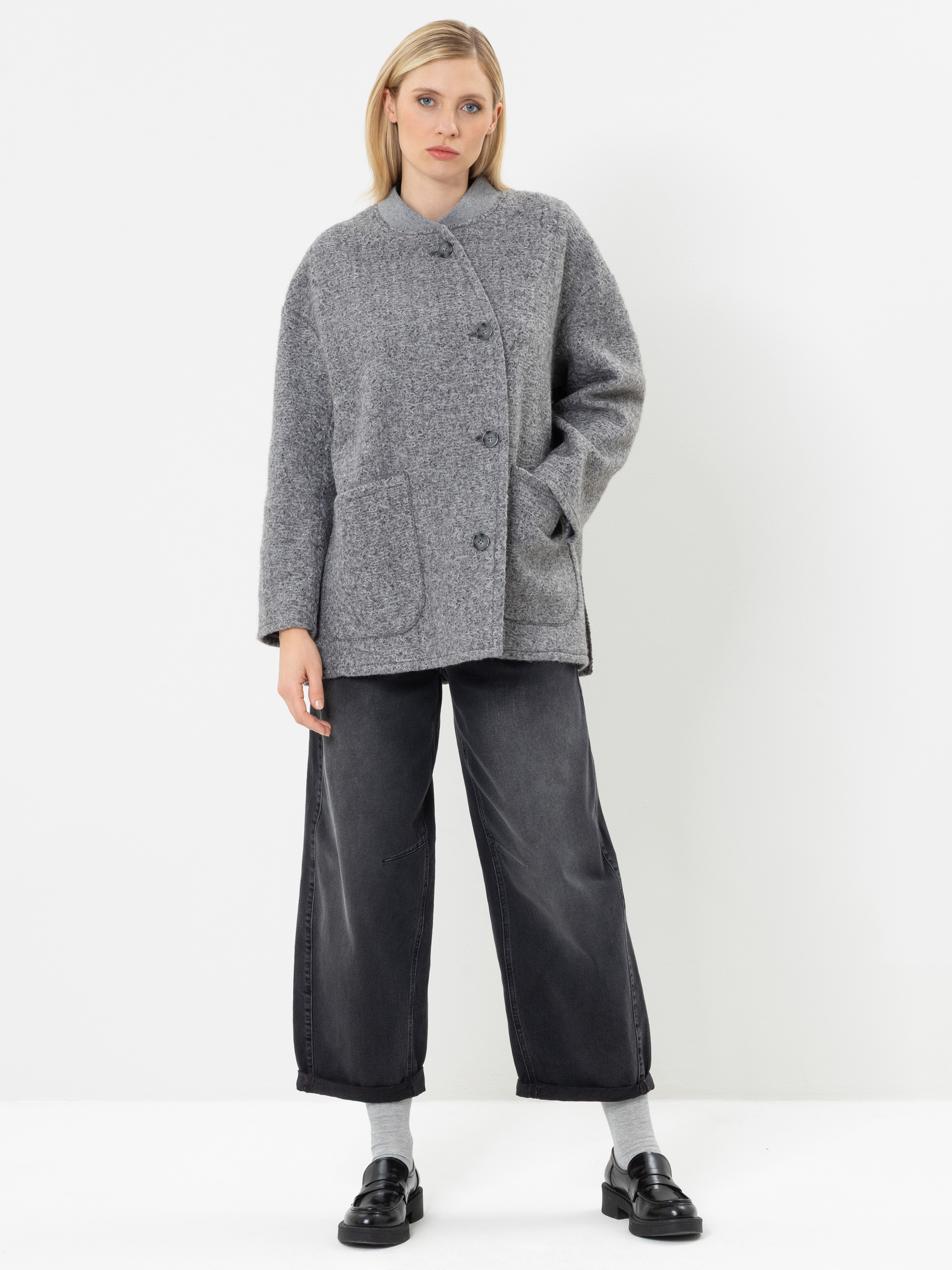 Eine Frau steht vor einem weißen Hintergrund und trägt einen grauen Mantel mit Knöpfen und großen Vordertaschen, eine schwarze Jeans mit weitem Bein, graue Socken und schwarze Slipper.