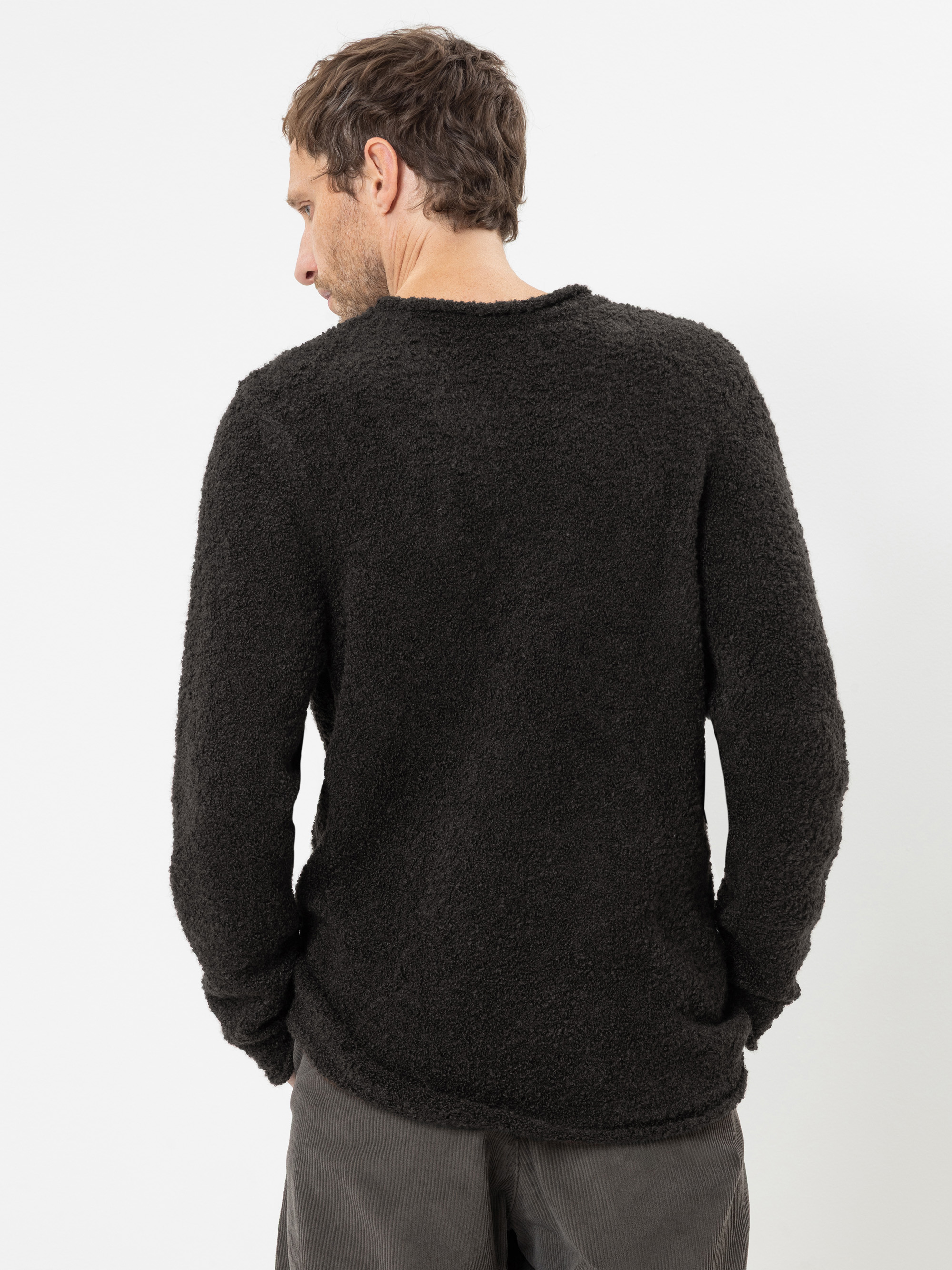 Ein Mann steht mit dem Rücken zur Kamera und trägt einen schwarzen, strukturierten Langarmpullover und eine graue Hose vor einem schlichten weißen Hintergrund.