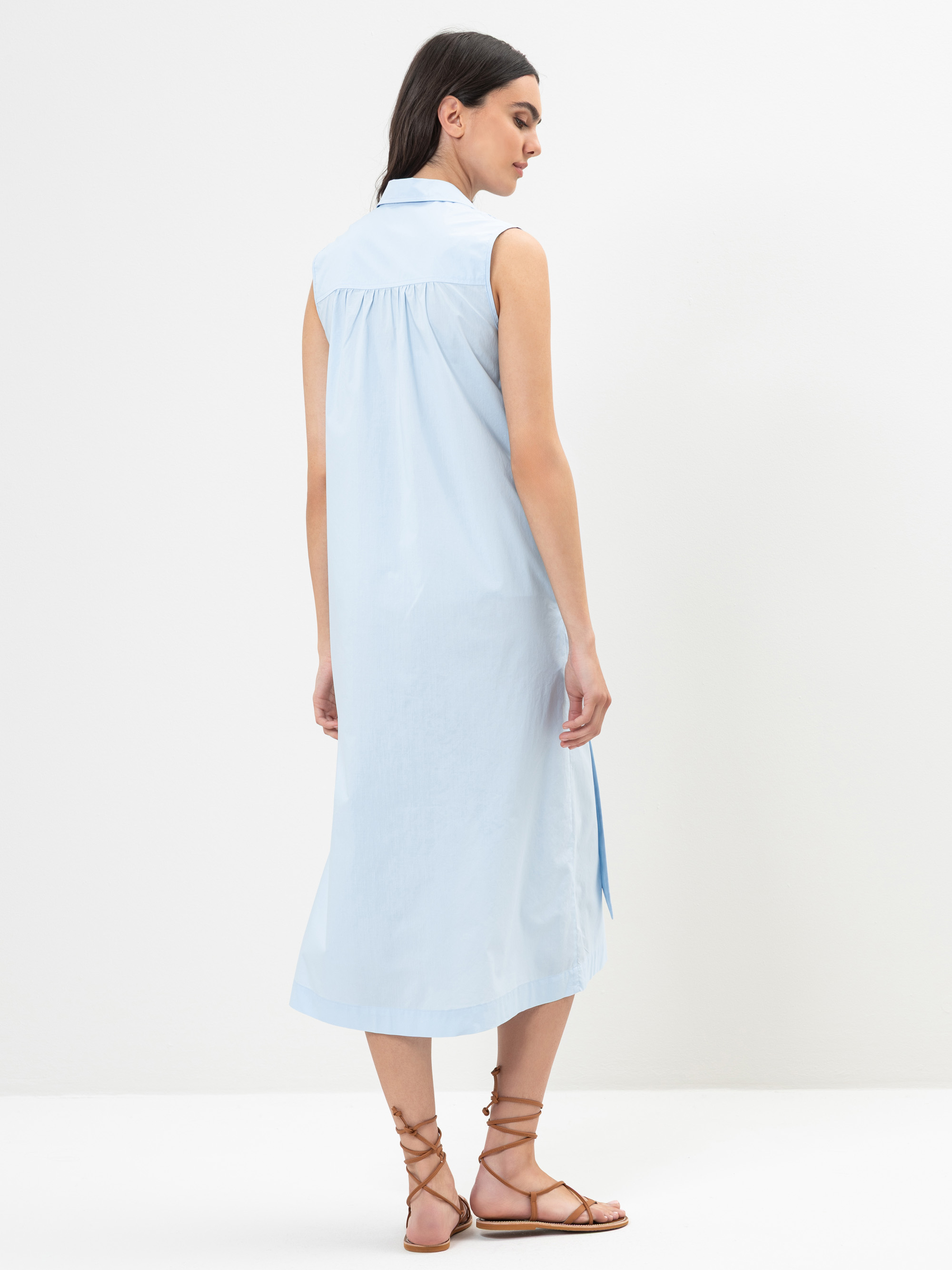 Eine Frau mit langen dunklen Haaren steht mit dem Gesicht zur Wand, trägt ein ärmelloses hellblaues Midi-Hemdkleid und braune Schnürsandalen vor einem schlichten weißen Hintergrund.