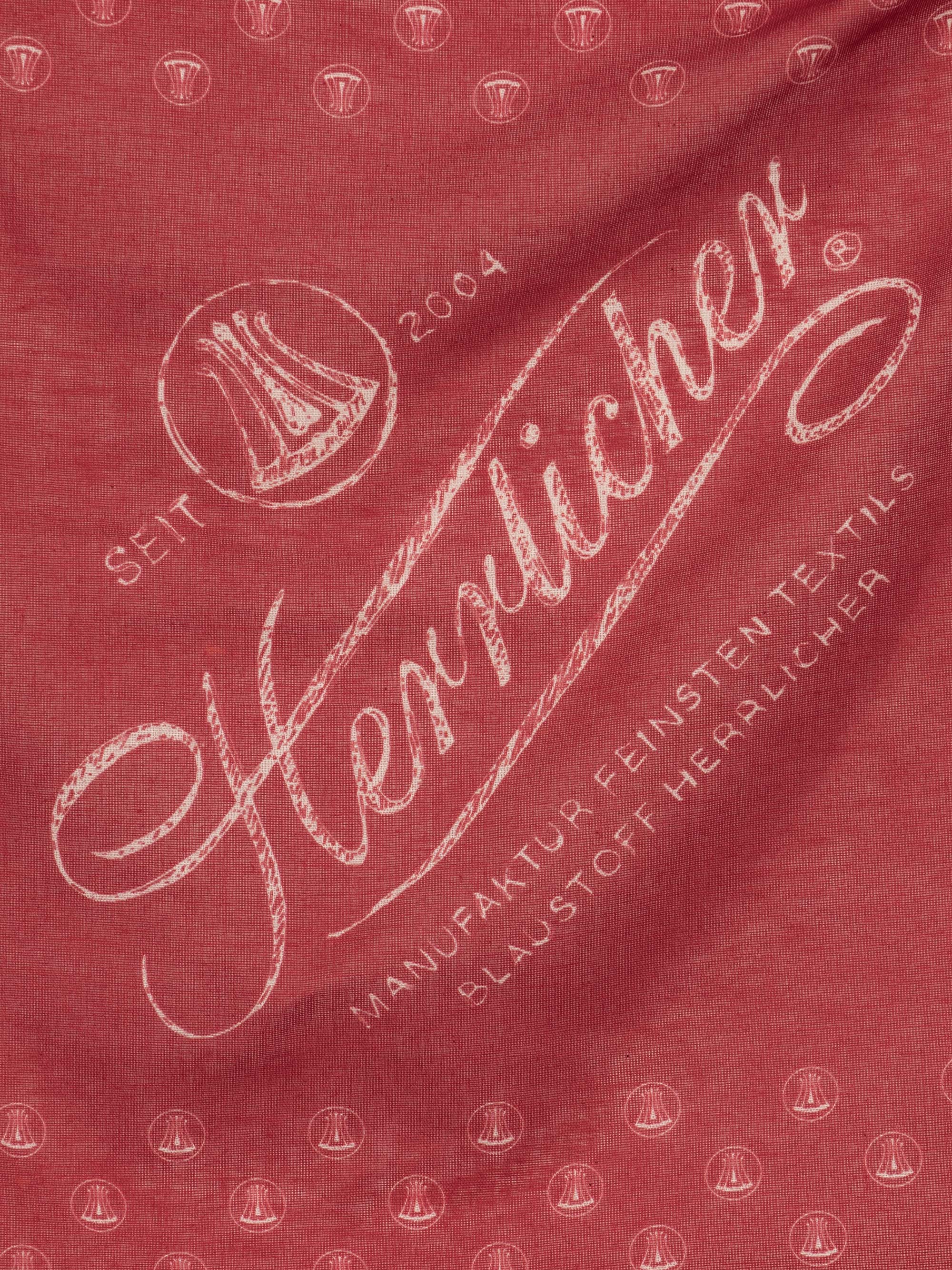 Roter Stoff mit weißer Schrift "Herrlicher" und Angaben wie "Seit 2004", "Manufaktur Feinster Textils" und "Blau Stoff Herrlicher" sowie kleinen kreisförmigen Logos mit einer stilisierten Figur.