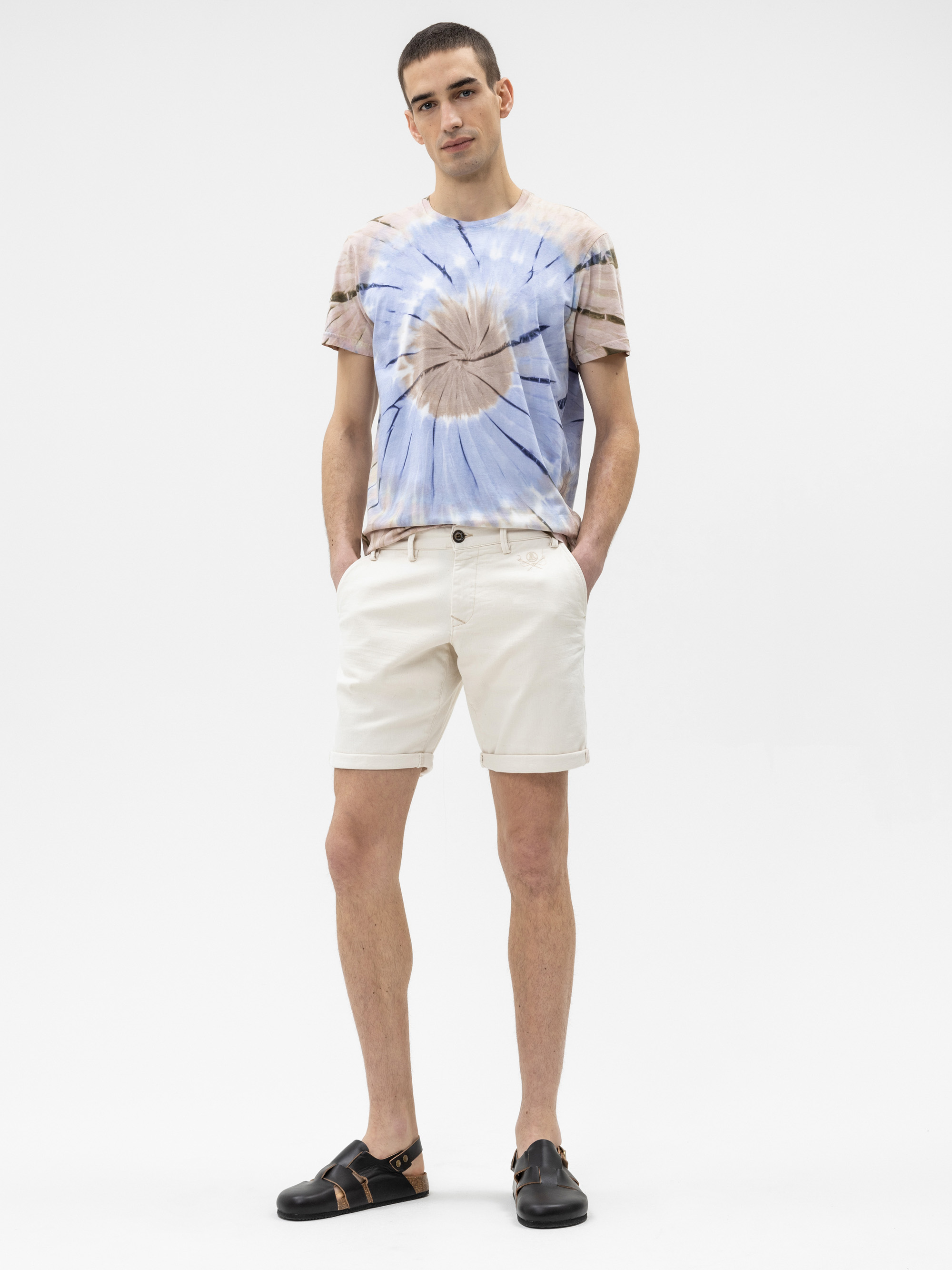 Eine Person steht vor einem einfarbig weißen Hintergrund und trägt ein tie-dye T-Shirt mit blauen, braunen und weißen Mustern, helle Shorts und schwarze Sandalen. Ihre Hände stecken in den Taschen.