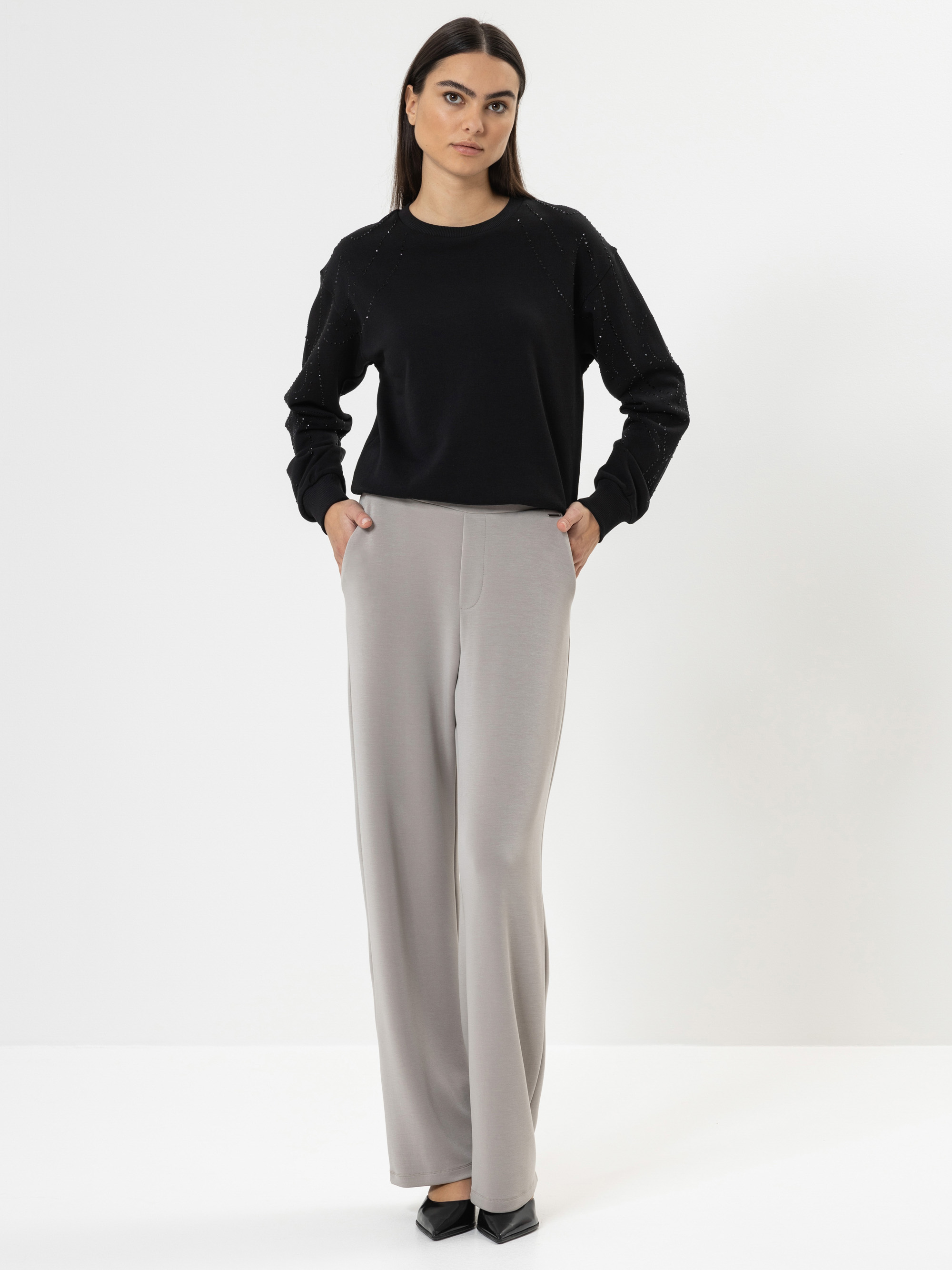 Eine Frau steht vor einem weißen Hintergrund, trägt ein schwarzes langärmeliges Oberteil mit dezenten Verzierungen und eine hellgraue Hose mit weitem Bein, die Hände in den Taschen und schwarze spitze Schuhe.