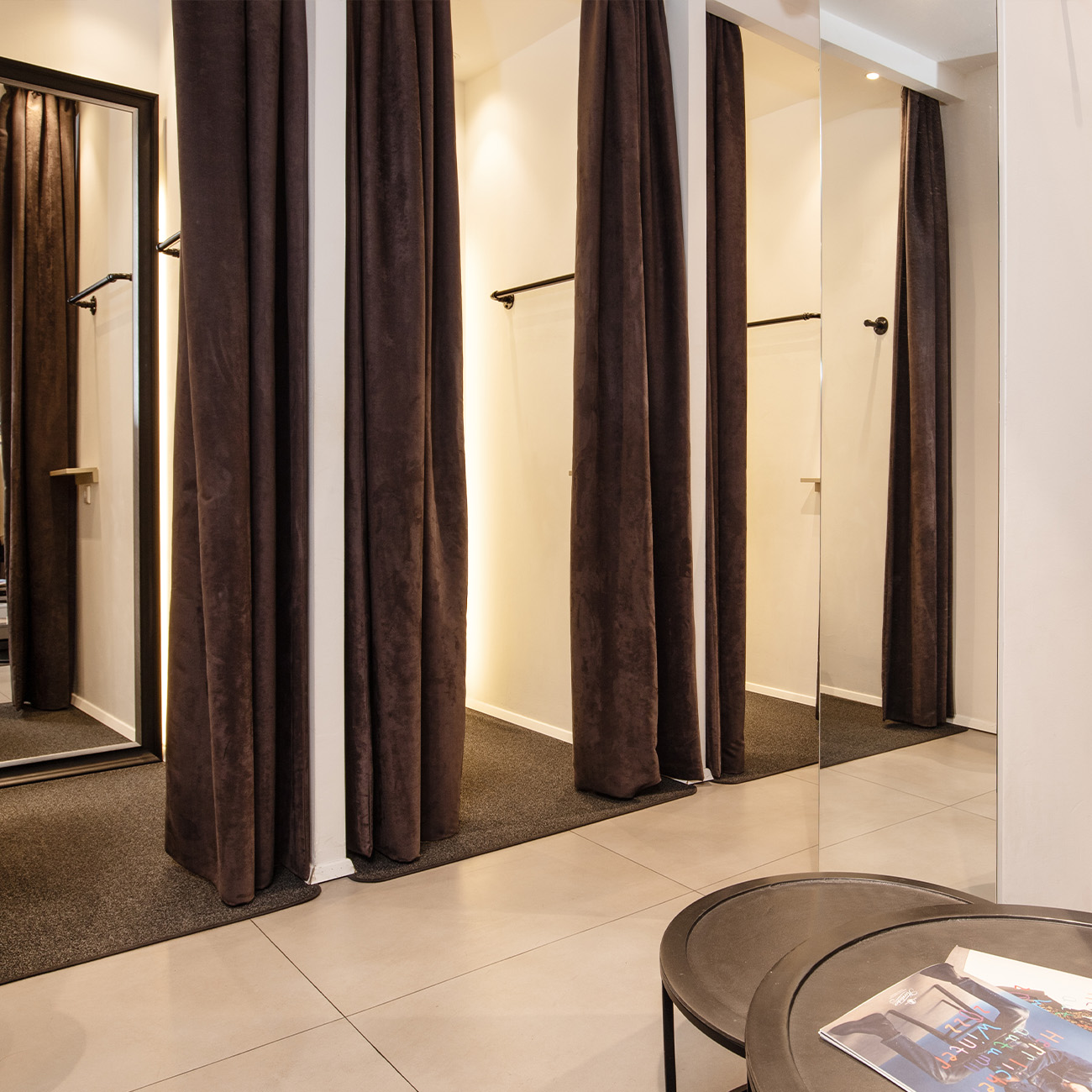 Eine Reihe von Umkleidekabinen mit dunklen Vorhängen in einem gut beleuchteten Raum; im Vordergrund ist ein runder Tisch mit Zeitschriften neben einem Wandspiegel zu sehen.