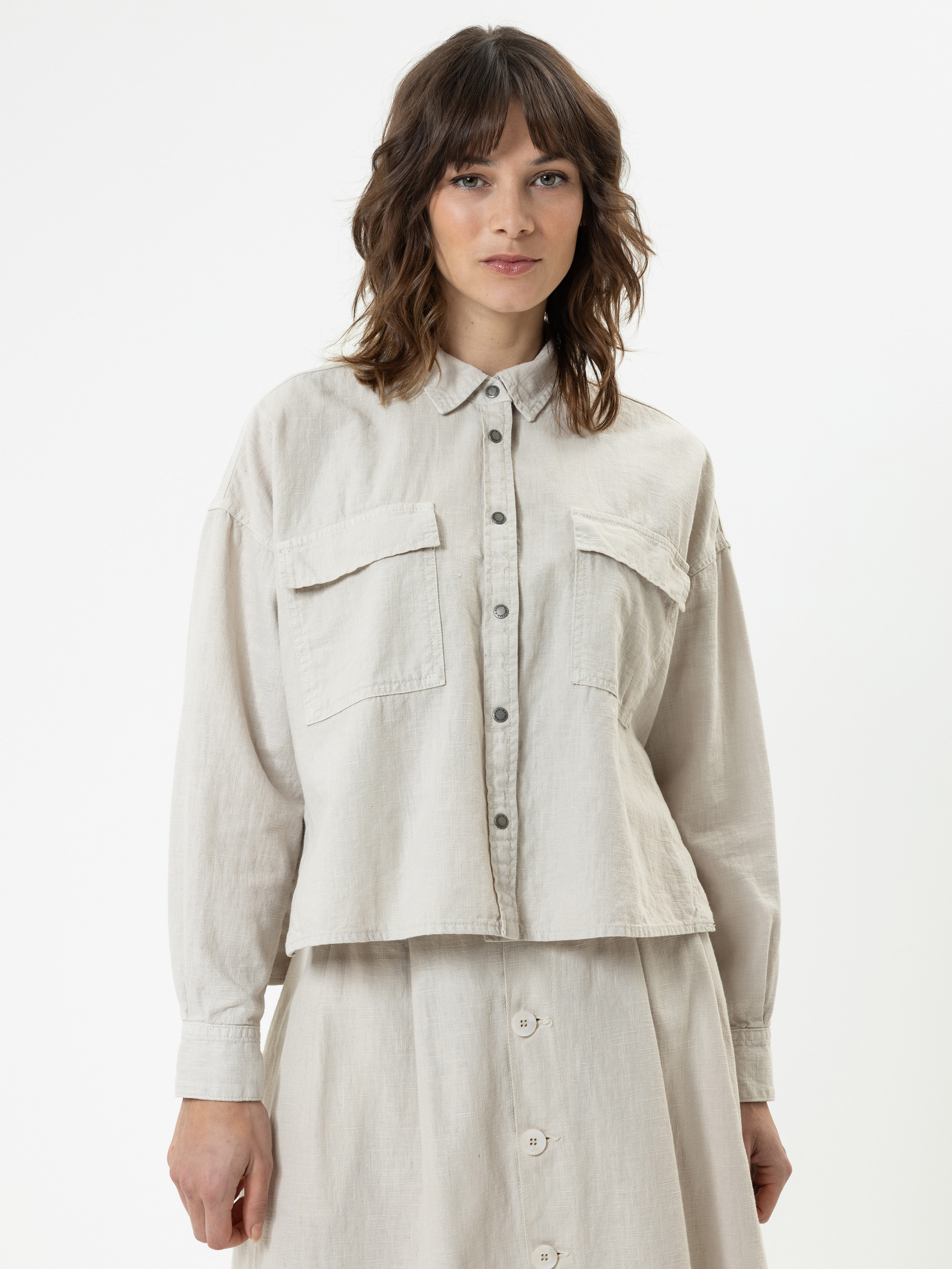 Eine Frau mit gewelltem braunem Haar trägt ein hellbeiges Button-up-Hemd mit Vordertaschen und einen passenden Rock. Sie steht mit dem Gesicht nach vorne vor einem schlichten weißen Hintergrund.