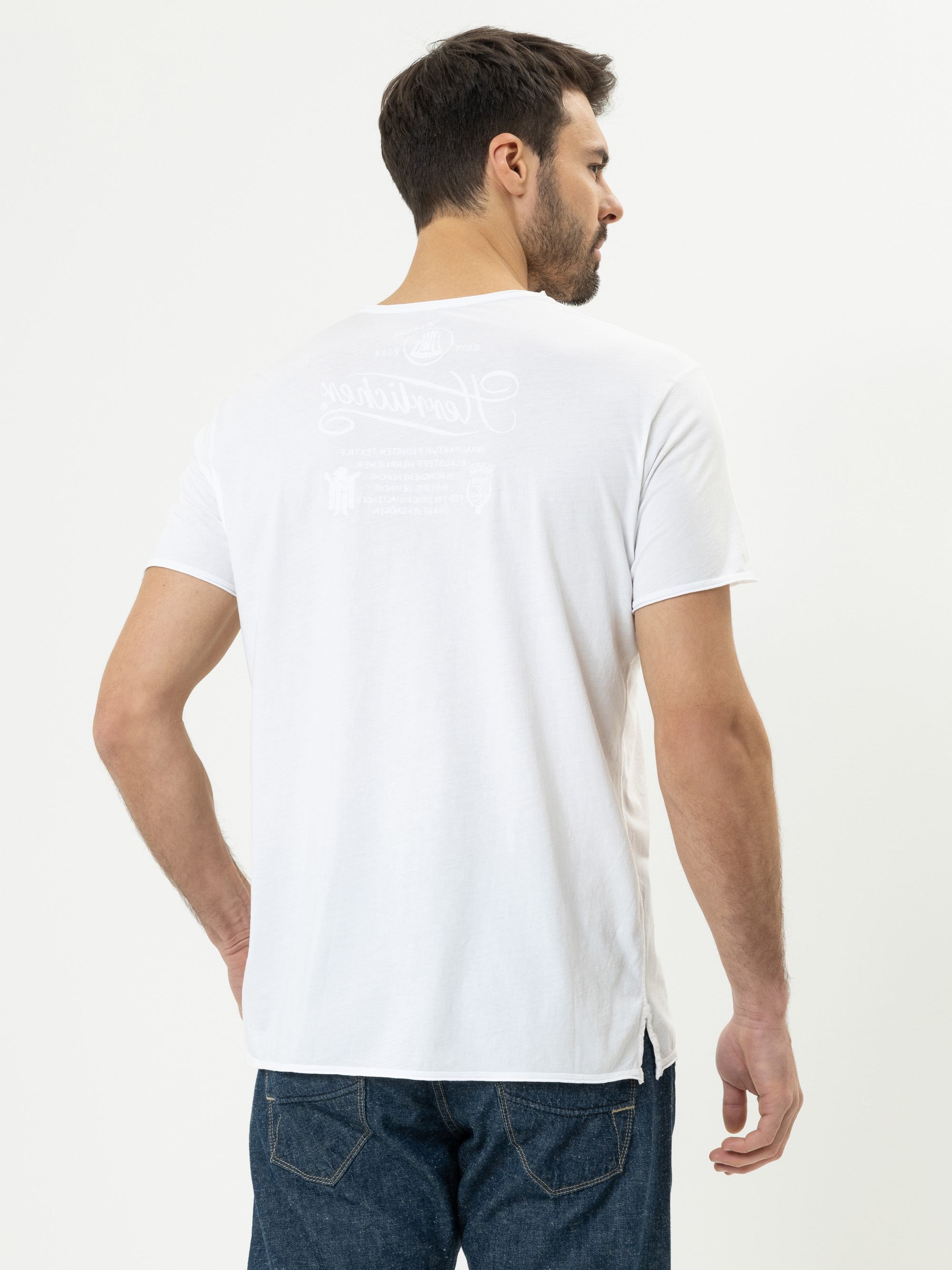 Ein Mann mit kurzen dunklen Haaren und einem Bart trägt ein schlichtes weißes T-Shirt und blaue Jeans. Er steht mit dem Rücken zur Kamera und blickt zur Seite vor einem weißen Hintergrund.