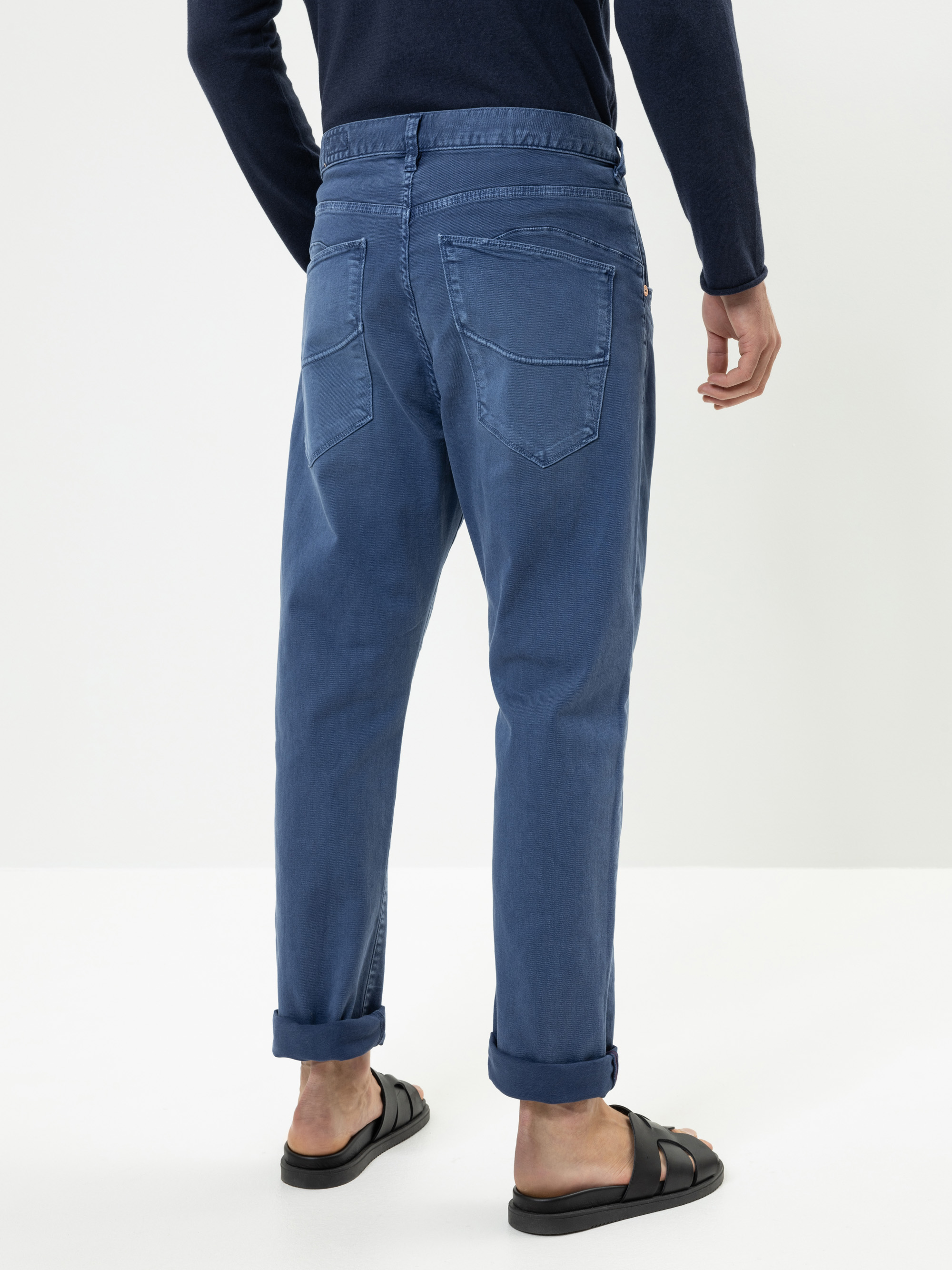 Rückansicht einer Person, die aufgerollte blaue Jeans, ein langärmeliges marineblaues Oberteil und schwarze Sandalen trägt und vor einem schlichten weißen Hintergrund steht.