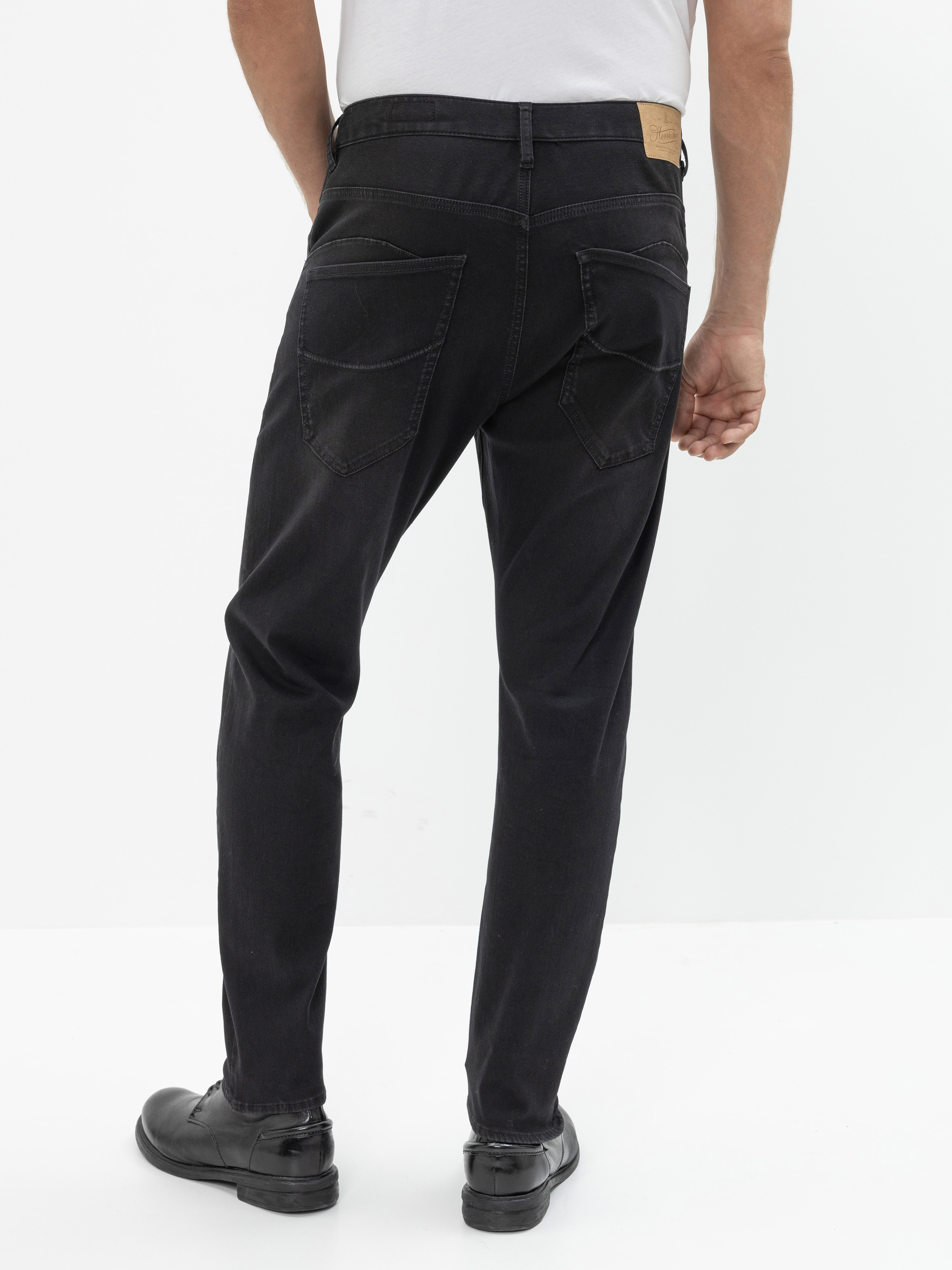 Mann in schwarzen Jeans und schwarzen Schuhen, von hinten gesehen, mit weißem Hemd, das in die Hose gesteckt ist. Der Hintergrund ist schlicht und weiß.