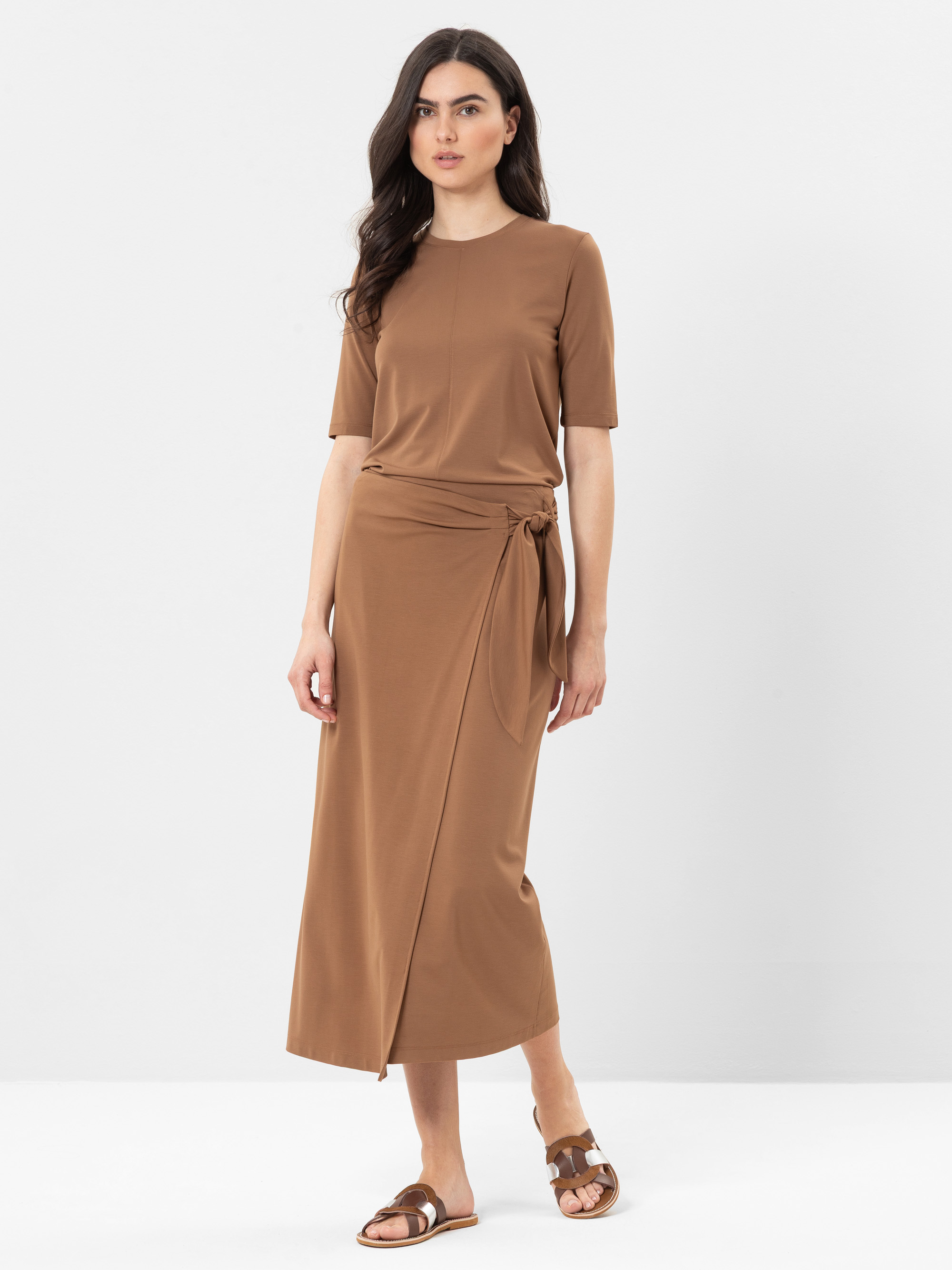 Eine Frau steht vor einem schlichten Hintergrund und trägt ein braunes, kurzärmeliges, knöchellanges Kleid mit einer seitlichen Bindung in der Taille und braune Sandalen. Ihr dunkles Haar ist offen und sie hat einen neutralen Gesichtsausdruck.