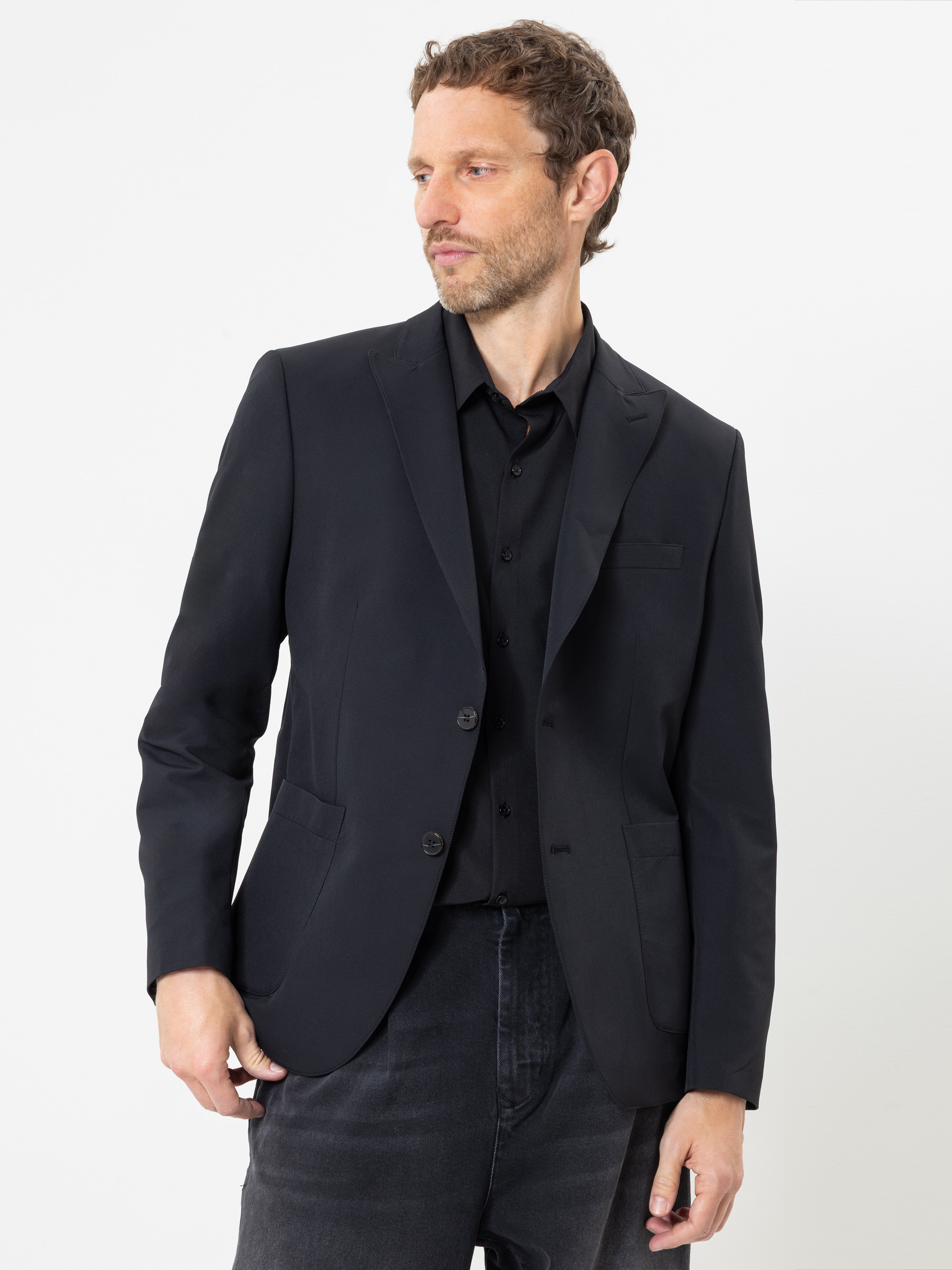 Ein Mann mit kurzen Locken, der ein schwarzes Sakko über einem schwarzen Hemd und einer schwarzen Jeans trägt, steht vor einem schlichten weißen Hintergrund und blickt nach links.