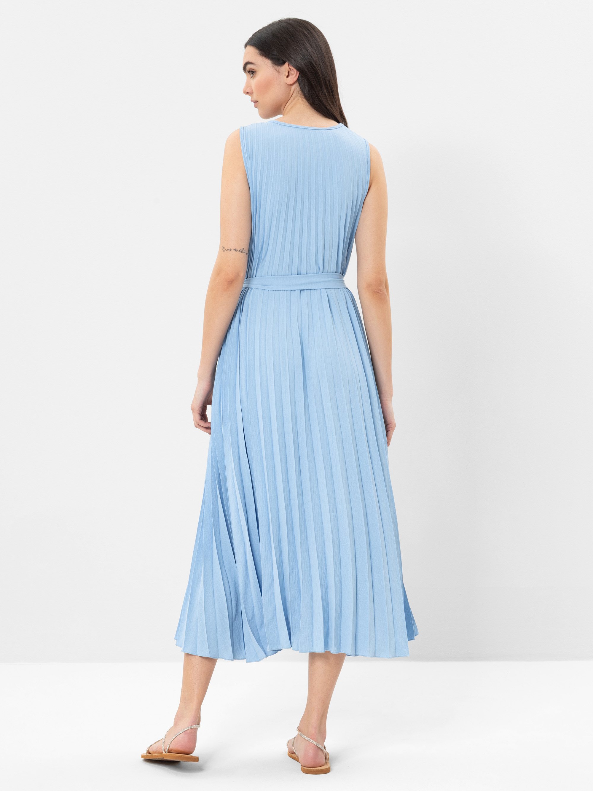 Eine Frau steht mit dem Gesicht nach hinten und trägt ein ärmelloses, hellblaues, plissiertes Midikleid mit einem Taillengürtel. Sie hat glattes dunkles Haar und trägt flache Sandalen. Der Hintergrund ist einfarbig weiß.