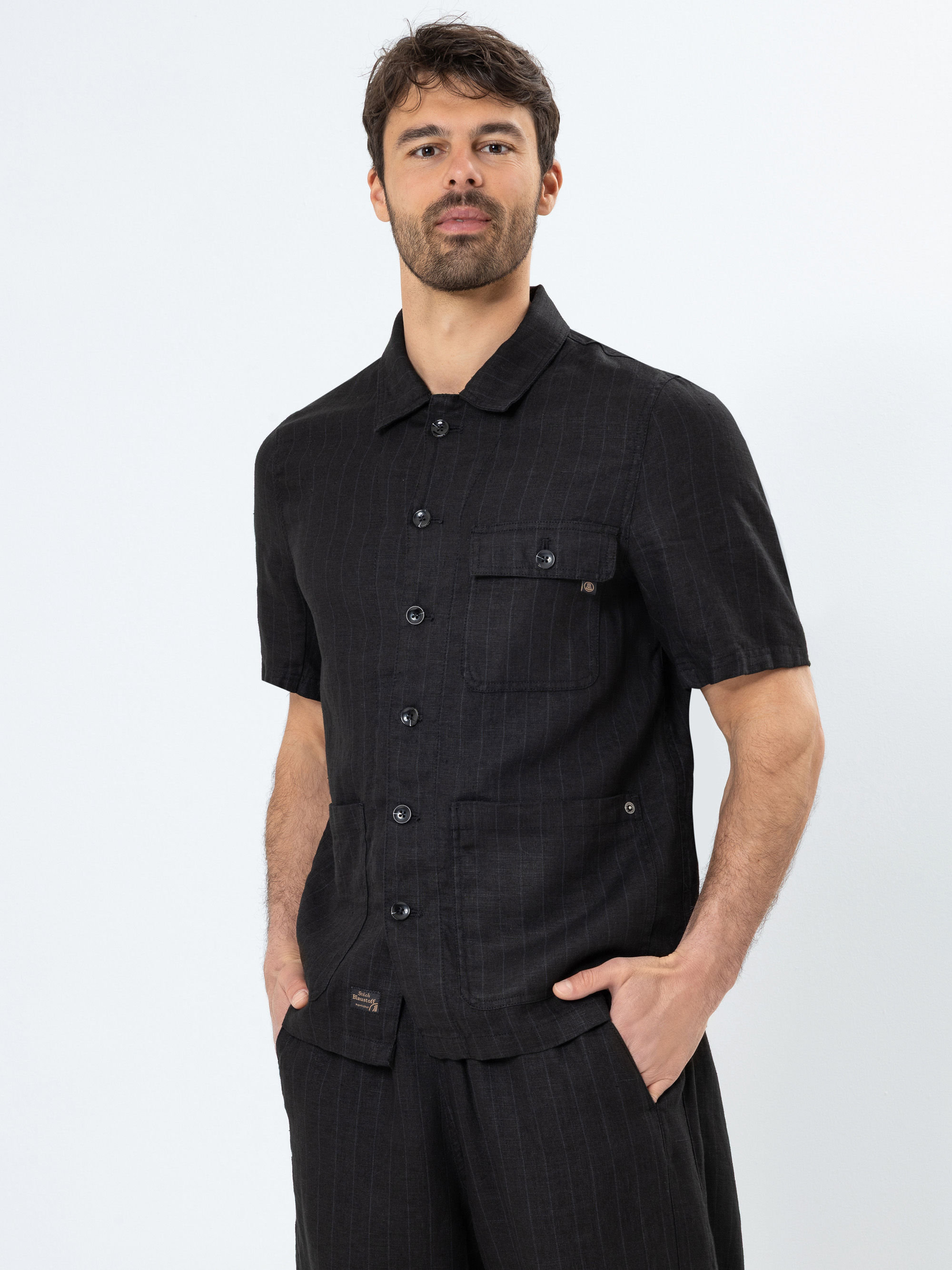 Ein Mann mit kurzen braunen Haaren und einem Bart trägt ein schwarzes, kurzärmeliges Button-up-Hemd mit Vordertaschen und eine passende Hose. Er steht vor einem schlichten weißen Hintergrund und hat die Hände in den Taschen.