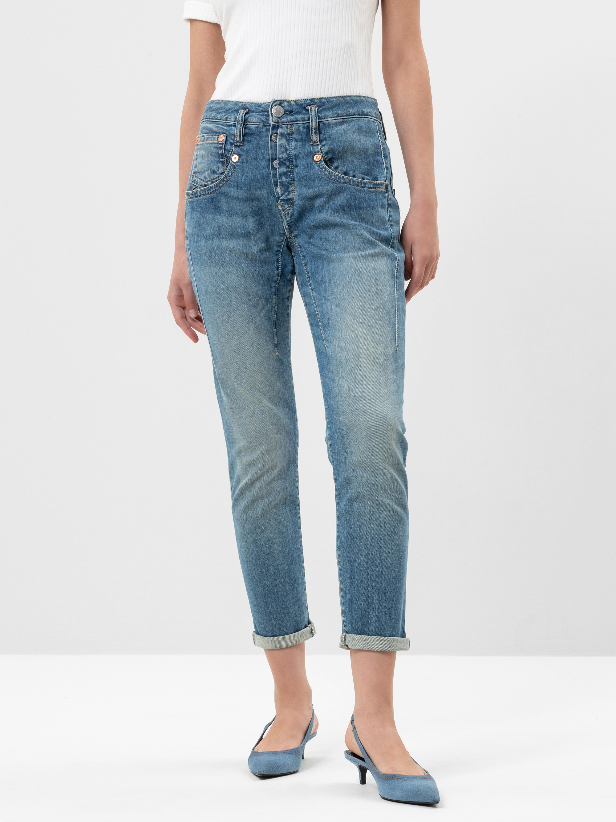 Eine Person, die hellblaue Jeans mit Bündchen, ein weißes geripptes Oberteil und blaue Halbschuhe trägt, steht vor einem schlichten weißen Hintergrund. Das Bild ist von den Schultern abwärts beschnitten.