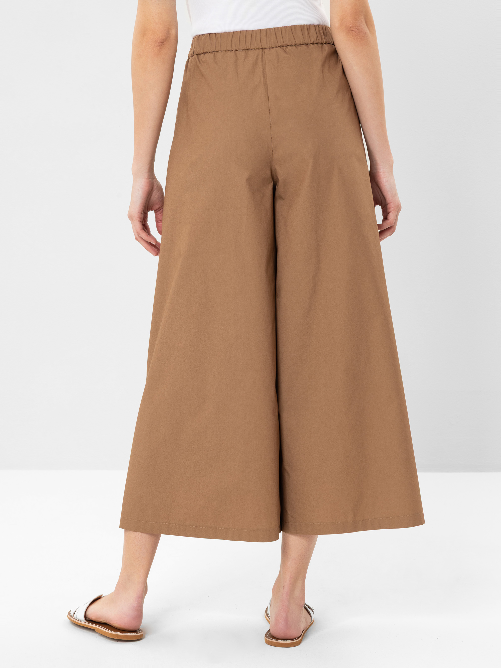 Eine Person, die eine weite, knöchellange braune Hose, ein weißes Oberteil und flache Sandalen trägt, wird von hinten vor einem schlichten hellen Hintergrund gezeigt.