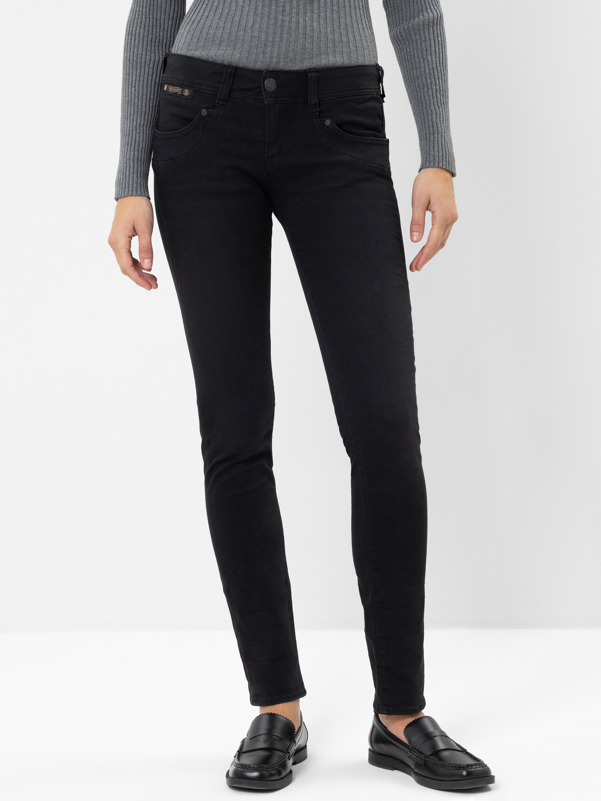 Eine Person, die ein graues, geripptes Langarmshirt, eine schwarze Skinny-Jeans und schwarze Slipper trägt, steht vor einem schlichten weißen Hintergrund. Das Bild ist von den Schultern abwärts beschnitten.