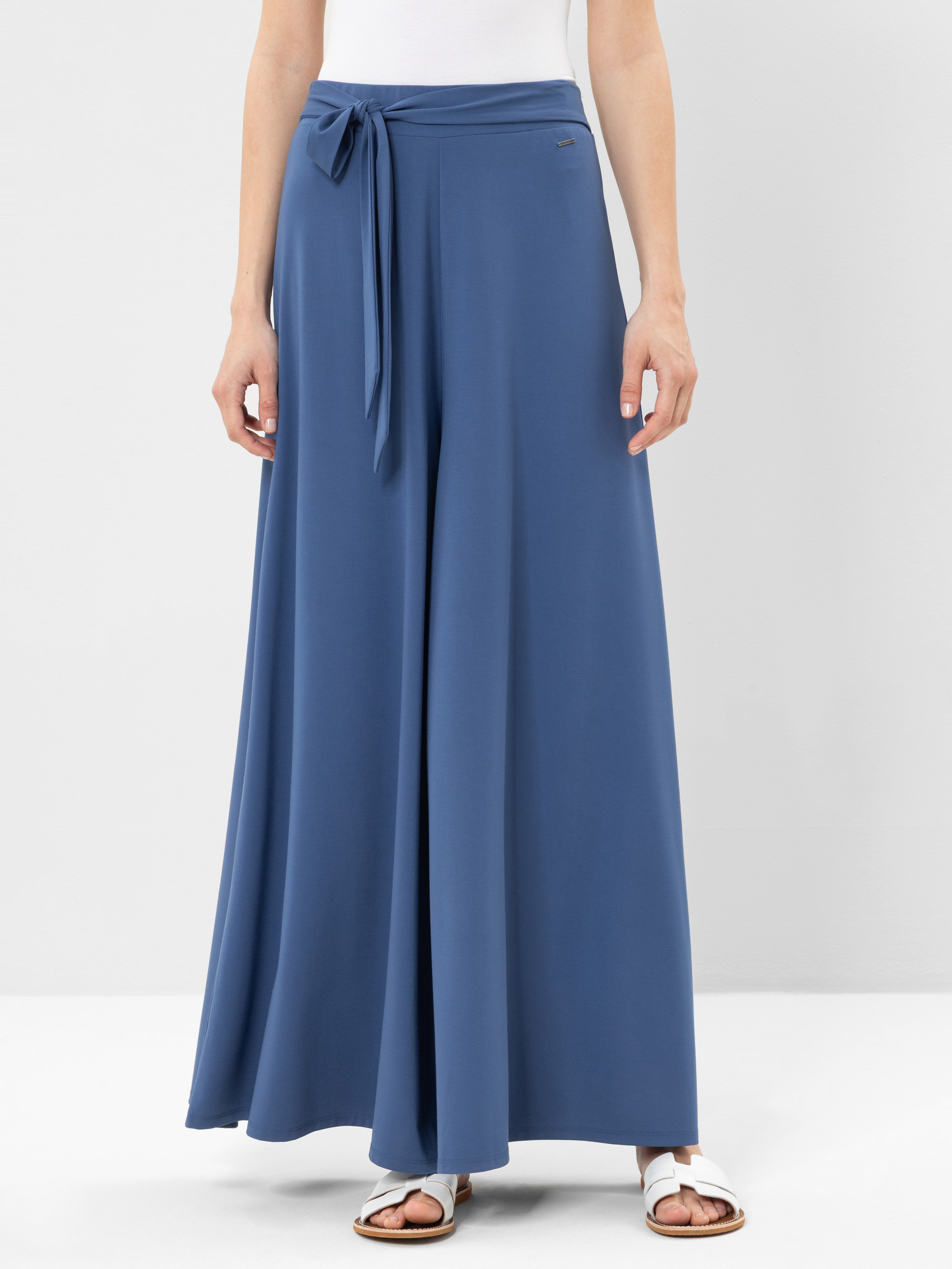 Eine Person, die eine blaue Palazzo-Hose mit hoher Taille und weitem Bein sowie einen passenden Stoffgürtel, ein weißes Oberteil und weiße flache Sandalen trägt, steht vor einem schlichten hellen Hintergrund.