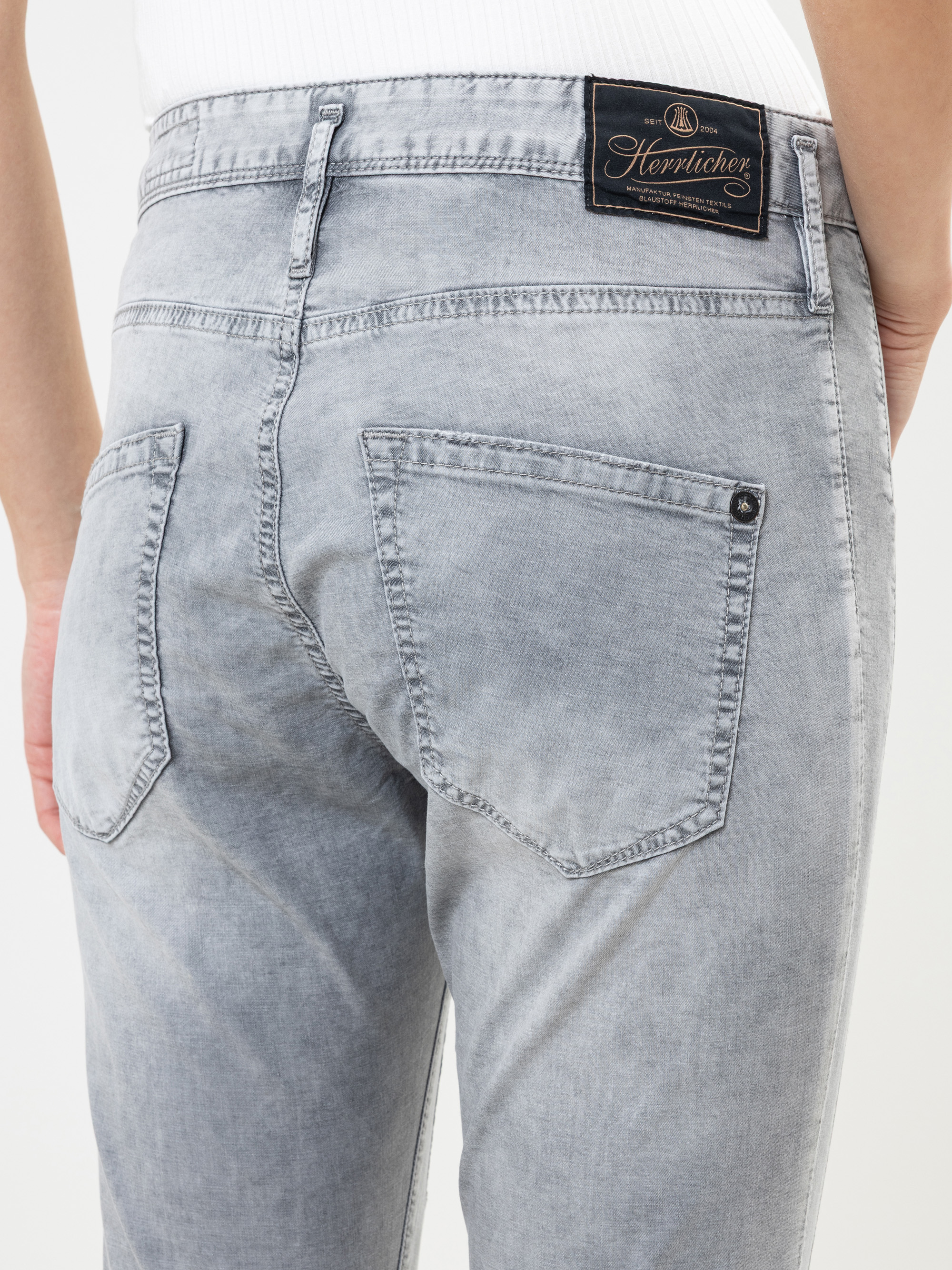 Nahaufnahme einer Person, die eine hellgraue Jeans mit zwei Gesäßtaschen und einem schwarzen Herrlicher-Markenetikett am Bund trägt. Die Person steht vor einem einfarbigen Hintergrund.