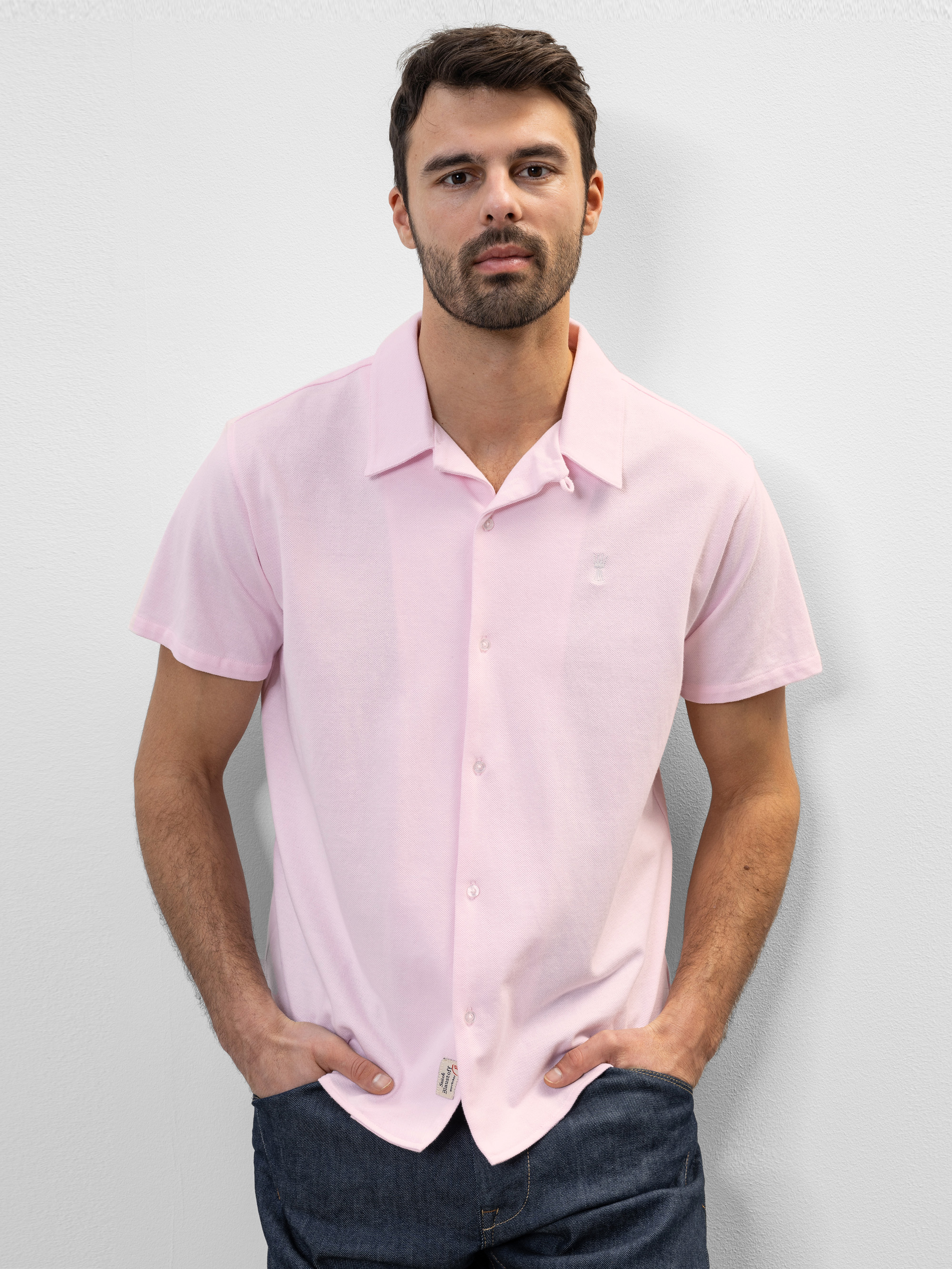Ein Mann mit kurzen dunklen Haaren und Bart trägt ein hellrosa, kurzärmeliges Button-up-Hemd und eine dunkle Hose und steht mit den Händen in den Taschen vor einem schlichten weißen Hintergrund.