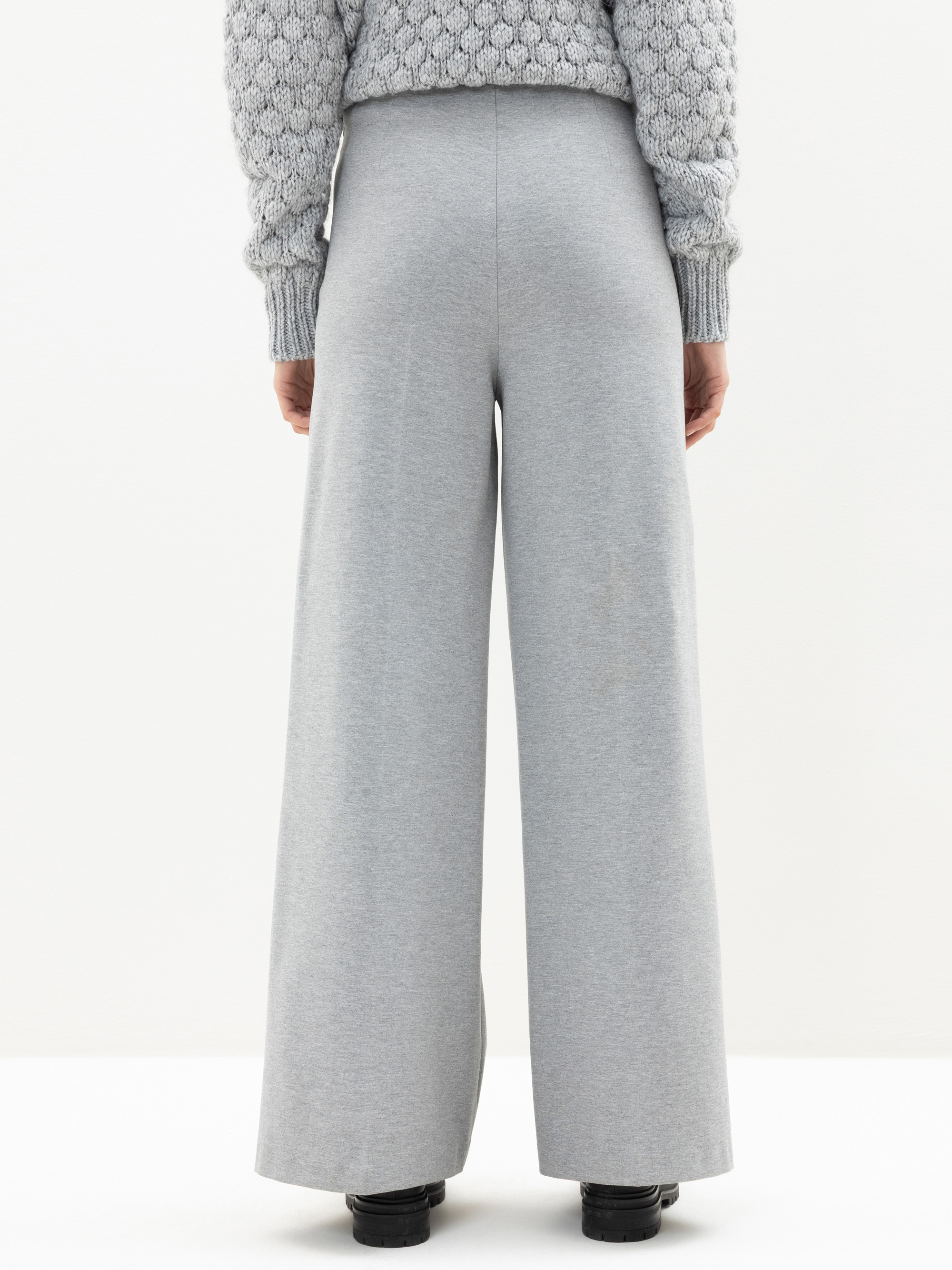 Person, die mit dem Rücken zur Kamera steht und einen strukturierten grauen Pullover und eine graue Hose mit hoher Taille und weitem Bein trägt. Das Outfit wird mit schwarzen Schuhen kombiniert, und der Hintergrund ist schlicht weiß.