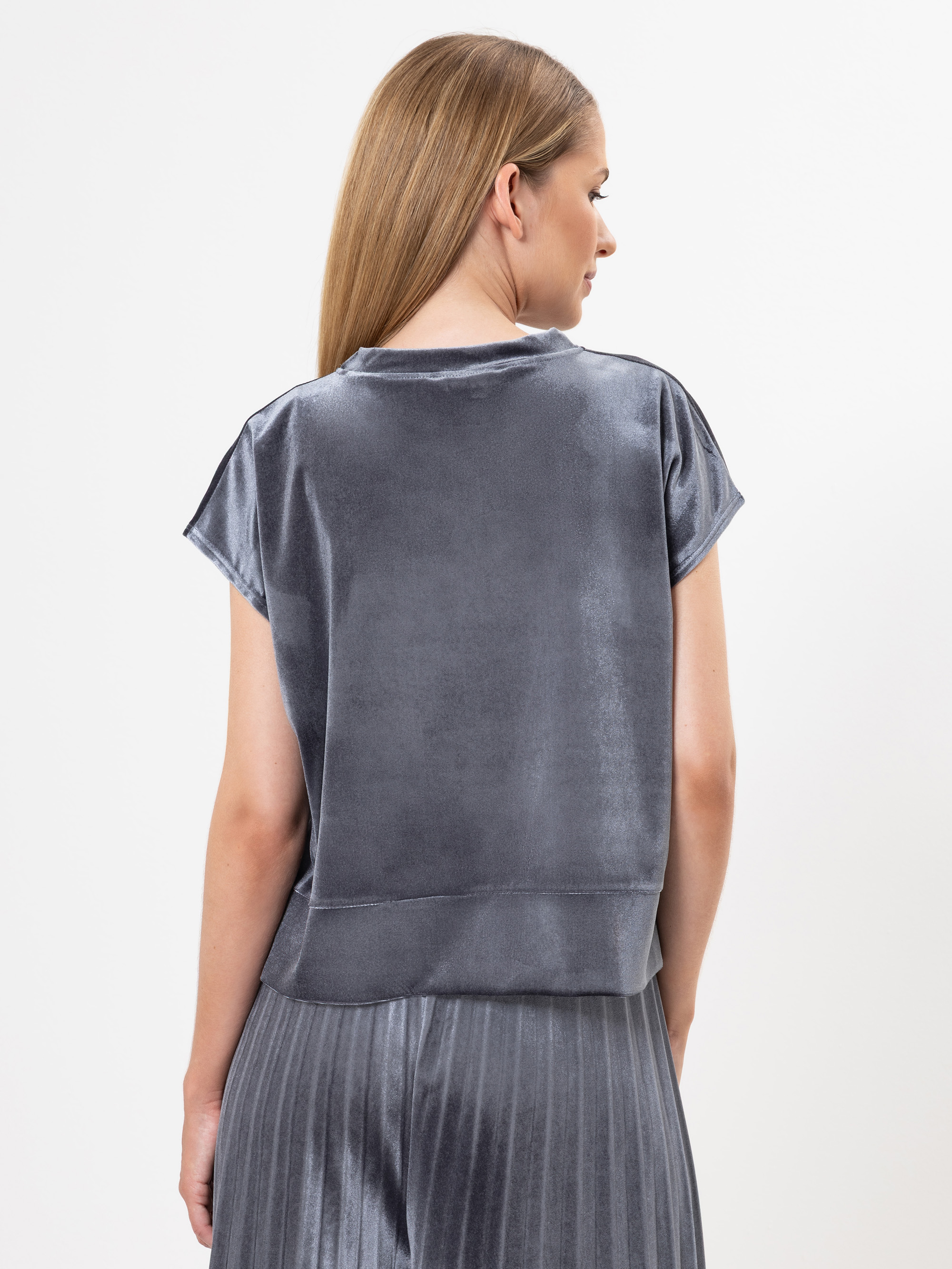 Eine Frau mit glattem, hellbraunem Haar trägt ein kurzärmeliges, locker sitzendes Oberteil aus metallgrauem Samt und einen dazu passenden Faltenrock, der von hinten vor einem schlichten weißen Hintergrund gezeigt wird.