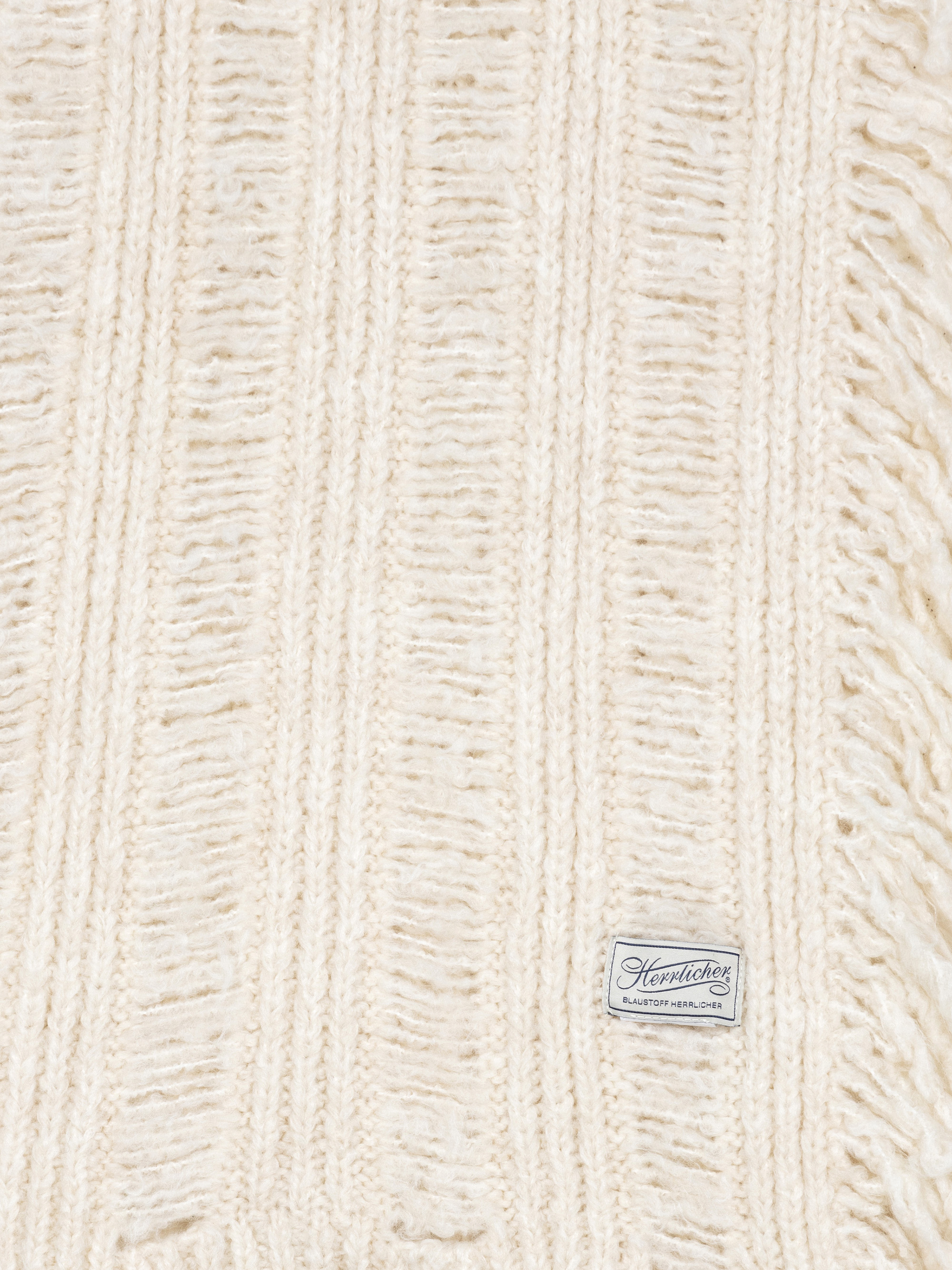Nahaufnahme eines cremefarbenen, gerippten Strickstoffs mit ausgefransten Rändern. Auf einem kleinen rechteckigen Etikett in der Ecke steht "Henriette Slip Soft Shoulder".