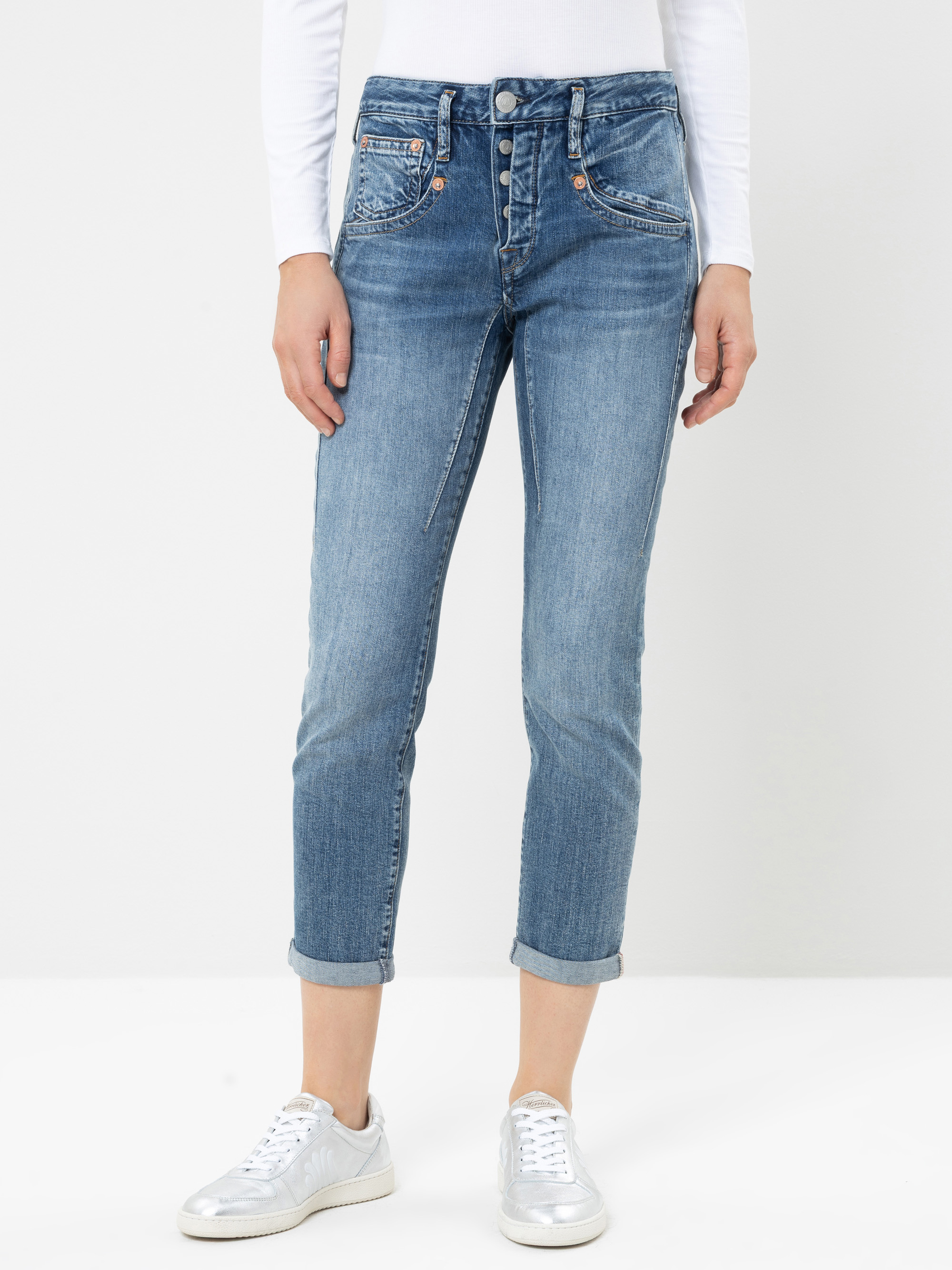 Eine Person, die hellblaue Jeans mit Umschlag, ein weißes Langarm-Oberteil und silberne Sneaker trägt, steht vor einem schlichten weißen Hintergrund. Das Bild zeigt die Person nur von den Schultern bis zu den Füßen.