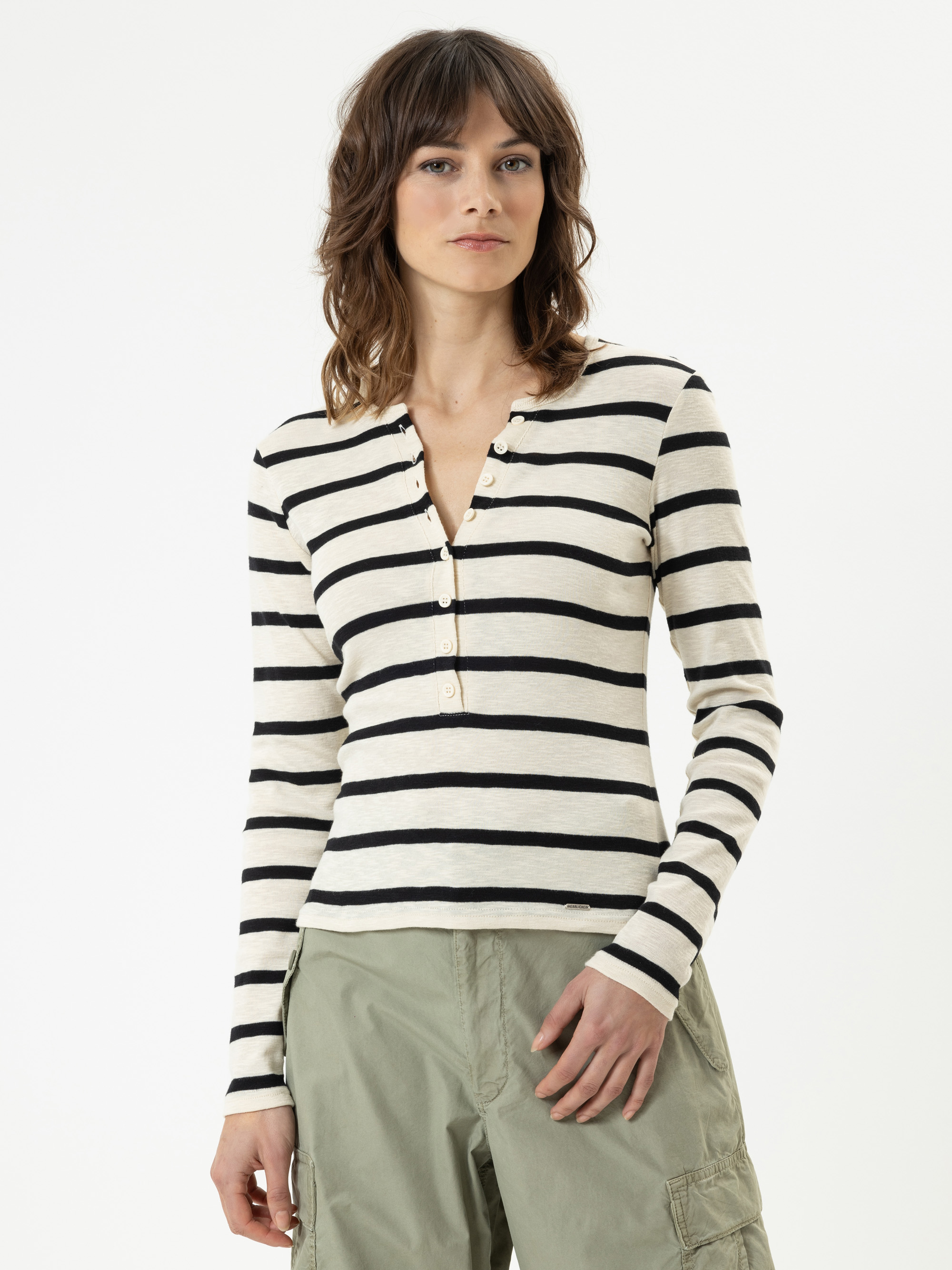 Eine Person mit gewelltem braunem Haar, die ein langärmeliges, cremefarben und schwarz gestreiftes Henley-Shirt und eine hellgrüne Cargohose trägt, posiert vor einem schlichten weißen Hintergrund.