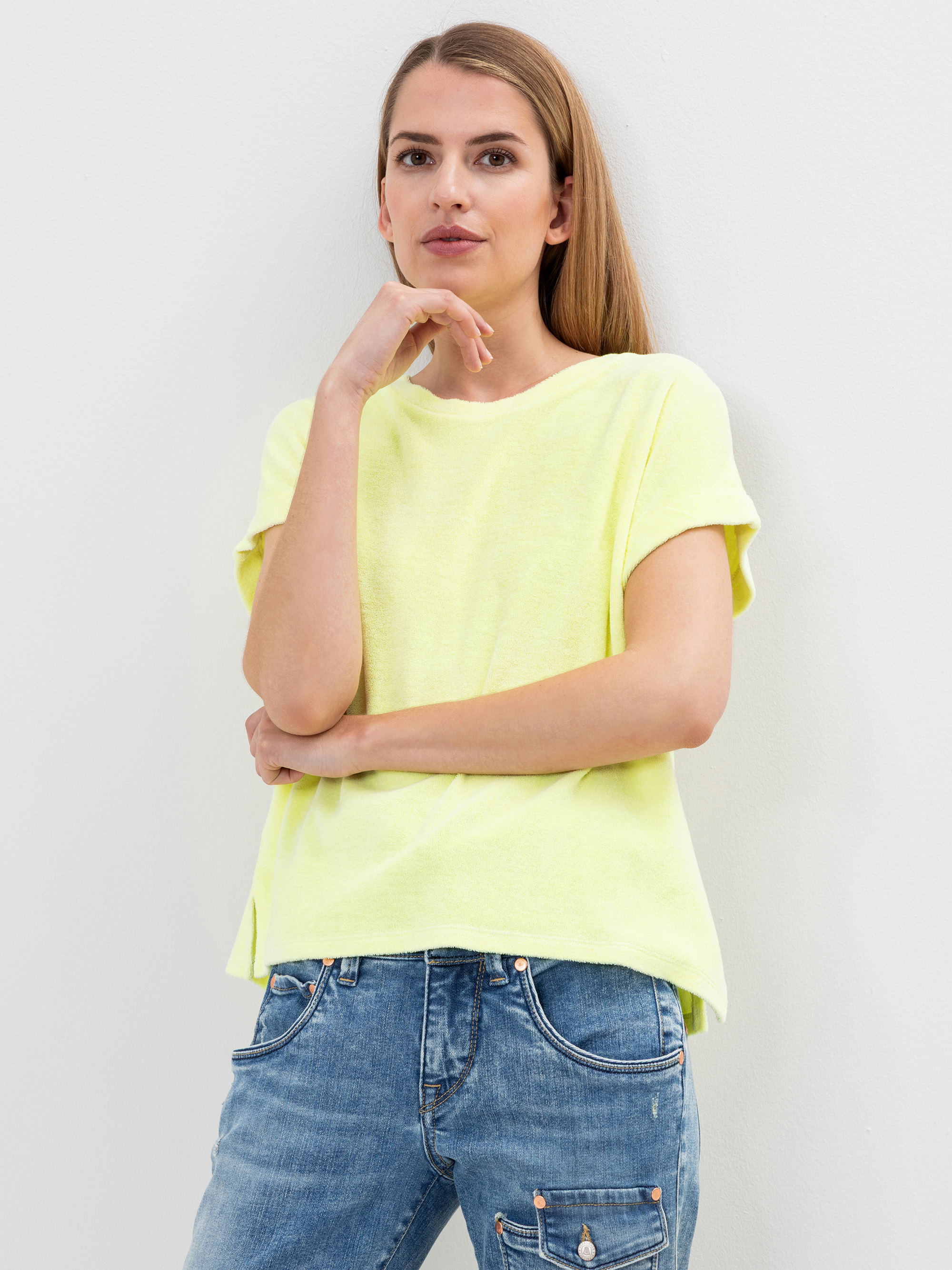 Eine Frau mit glattem, hellbraunem Haar trägt ein lockeres, kurzärmeliges gelbes Oberteil und blaue Jeans. Sie steht vor einer schlichten weißen Wand, stützt ihre Hand auf ihr Kinn und blickt mit einem leichten Lächeln in die Kamera.