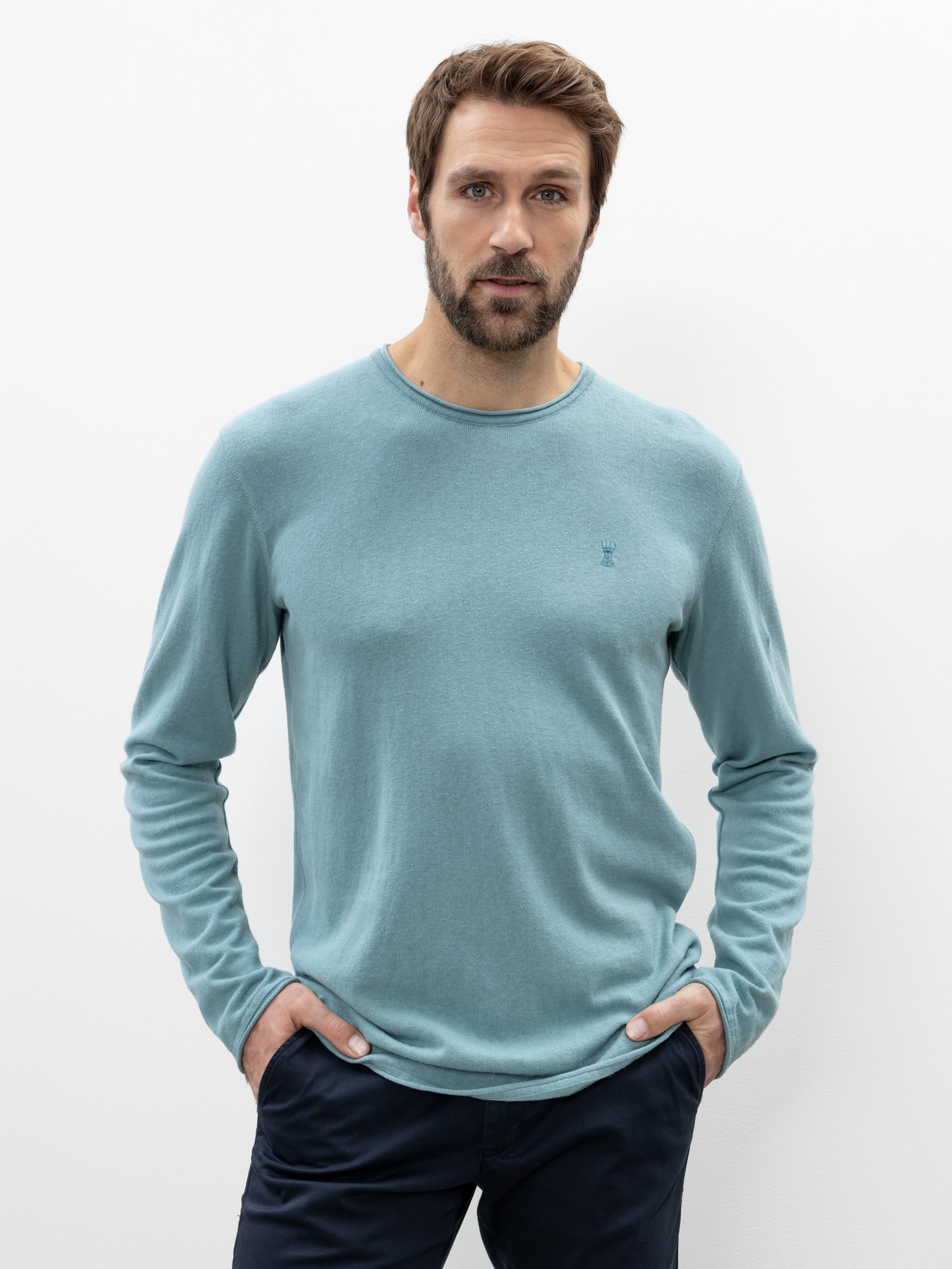 Ein Mann mit hellbraunem Haar und Bart trägt ein hellblaues langärmeliges Hemd und eine dunkle Hose und steht mit den Händen in den Taschen vor einem schlichten weißen Hintergrund.