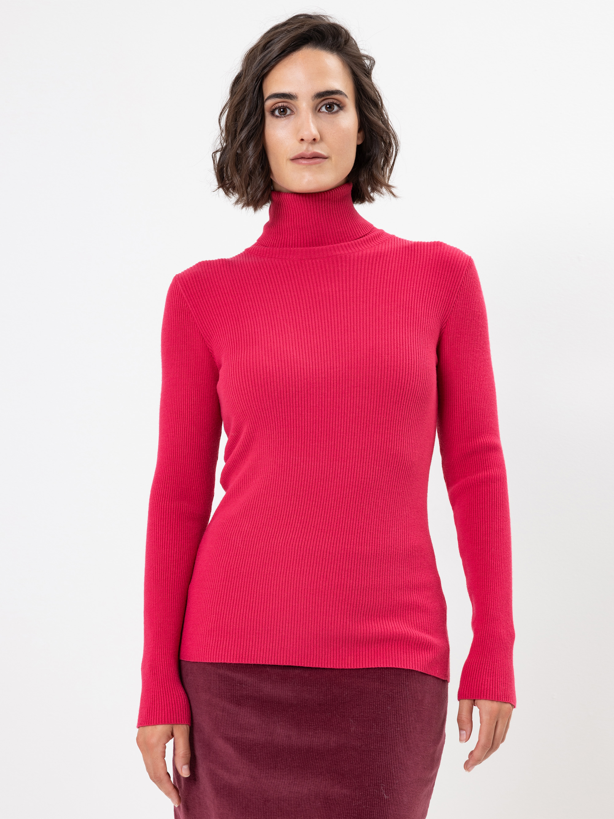 Eine Frau mit kurzen braunen Haaren trägt einen hellrosa Rollkragenpullover und einen dunkelrosa Rock und steht vor einem schlichten weißen Hintergrund. Sie blickt nach vorne und hat einen neutralen Gesichtsausdruck.