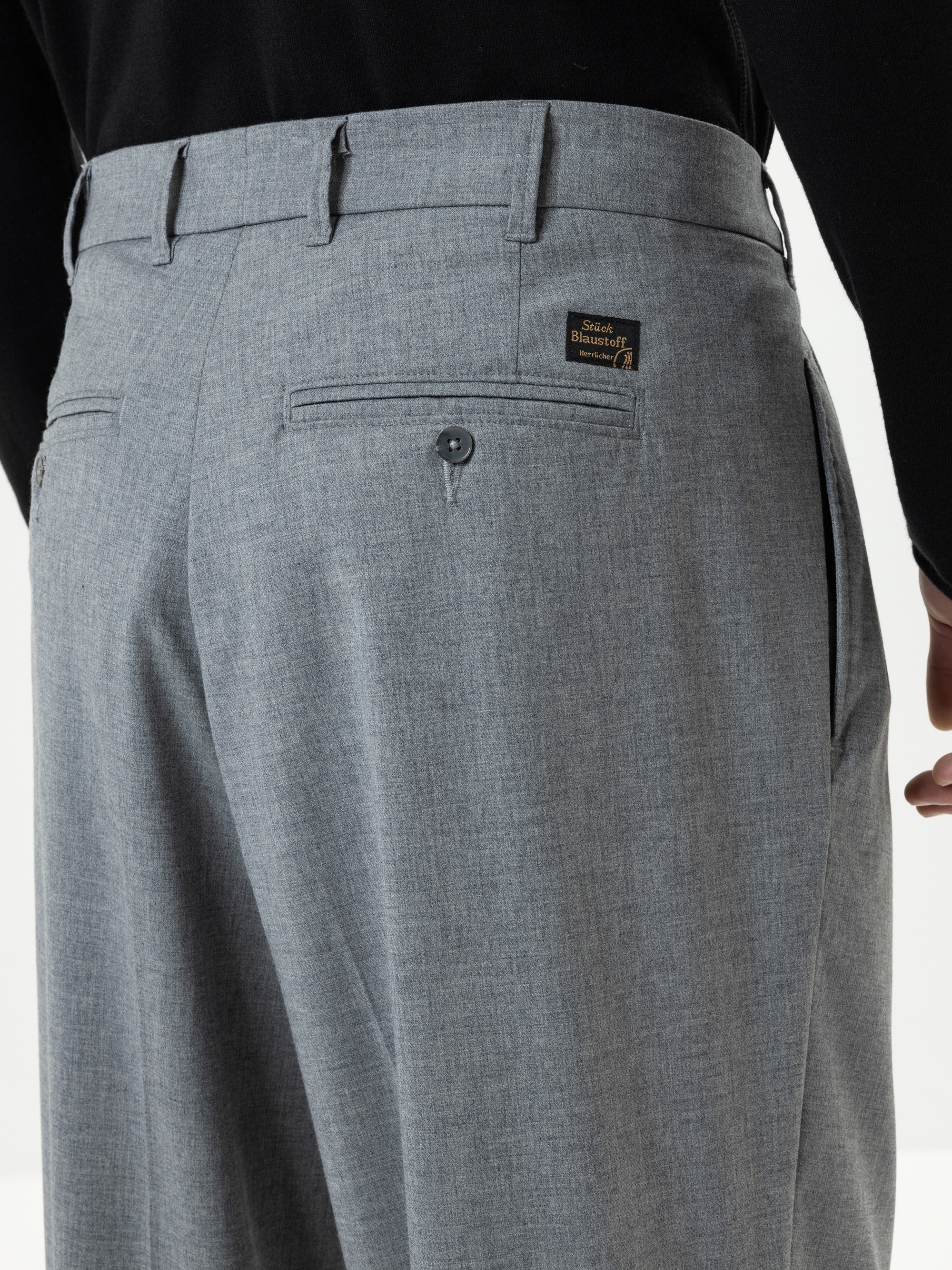 Nahaufnahme einer Person, die eine graue Anzughose und ein schwarzes Oberteil trägt, wobei der Schwerpunkt auf dem Bereich der Gesäßtasche liegt. Über der Tasche ist ein kleines schwarz-goldenes Etikett mit der Aufschrift Sustainable Business Attire z