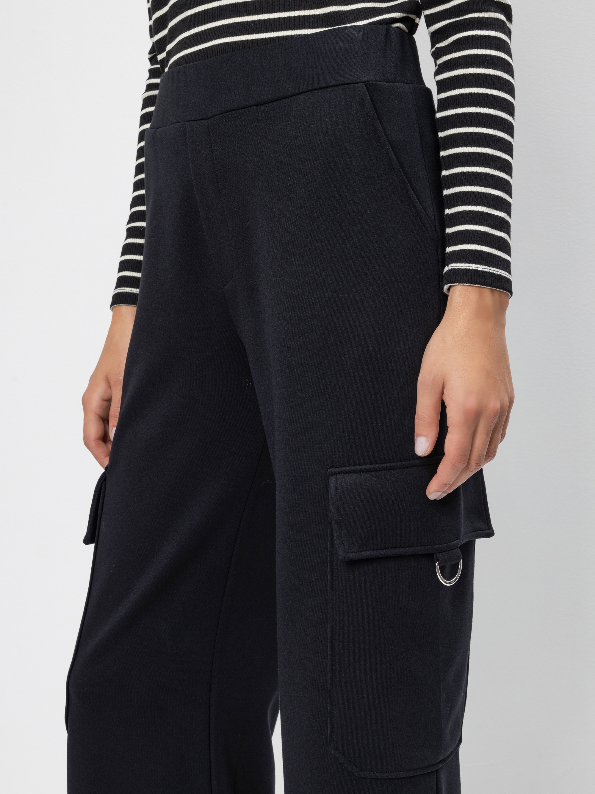 Eine Person trägt eine schwarze Cargohose mit Seitentaschen und ein schwarz-weiß gestreiftes langärmeliges Oberteil, das von der Mitte der Brust bis zur Mitte der Oberschenkel vor einem unifarbenen Hintergrund gezeigt wird.