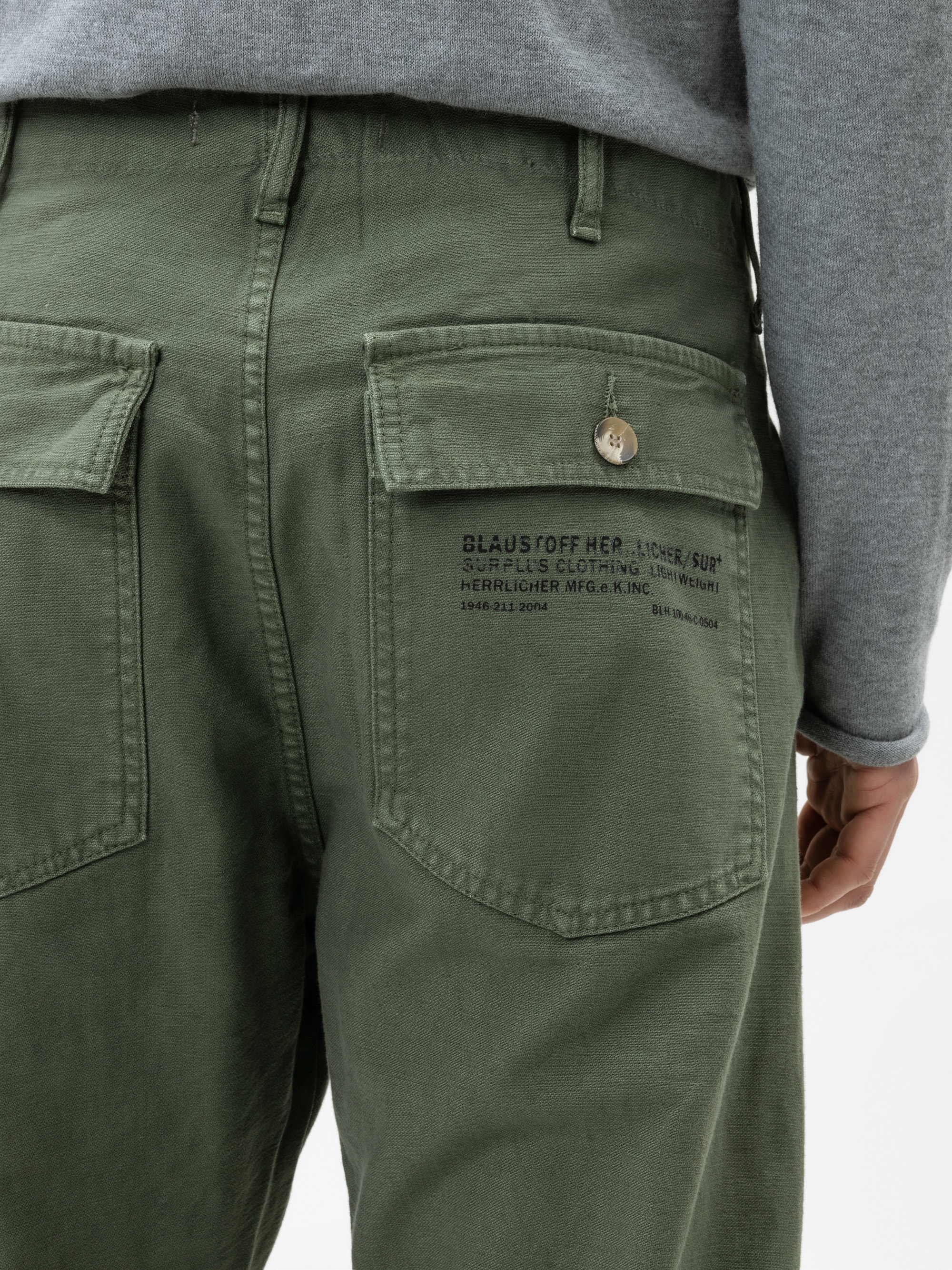 Nahaufnahme des Rückens einer Person, die eine olivgrüne Hose mit großen Klappentaschen und schwarzem Aufdruck auf einer Tasche trägt. Die Person trägt außerdem ein graues langärmeliges Hemd.