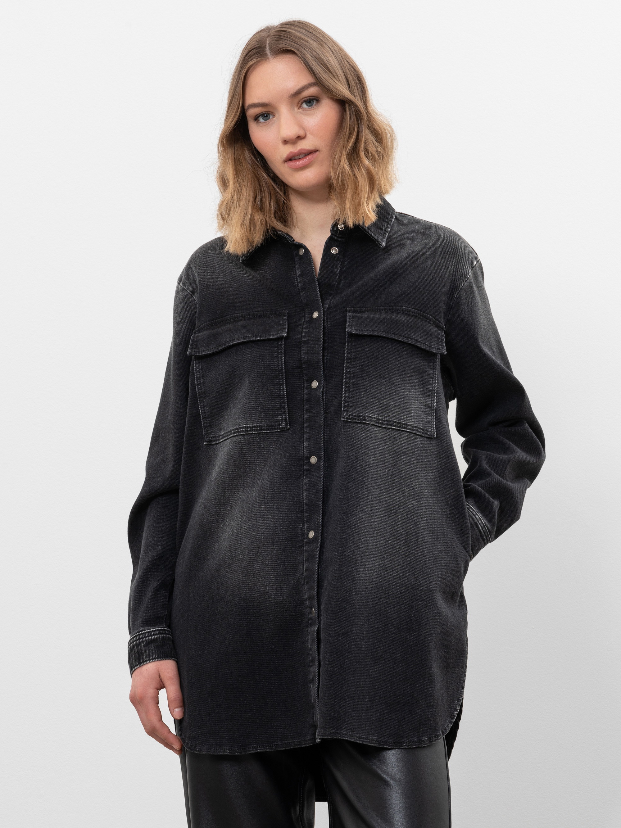 Eine Frau mit schulterlangem hellbraunem Haar trägt ein locker sitzendes schwarzes Jeanshemd mit Vordertaschen und eine schwarze Hose vor einem schlichten weißen Hintergrund.