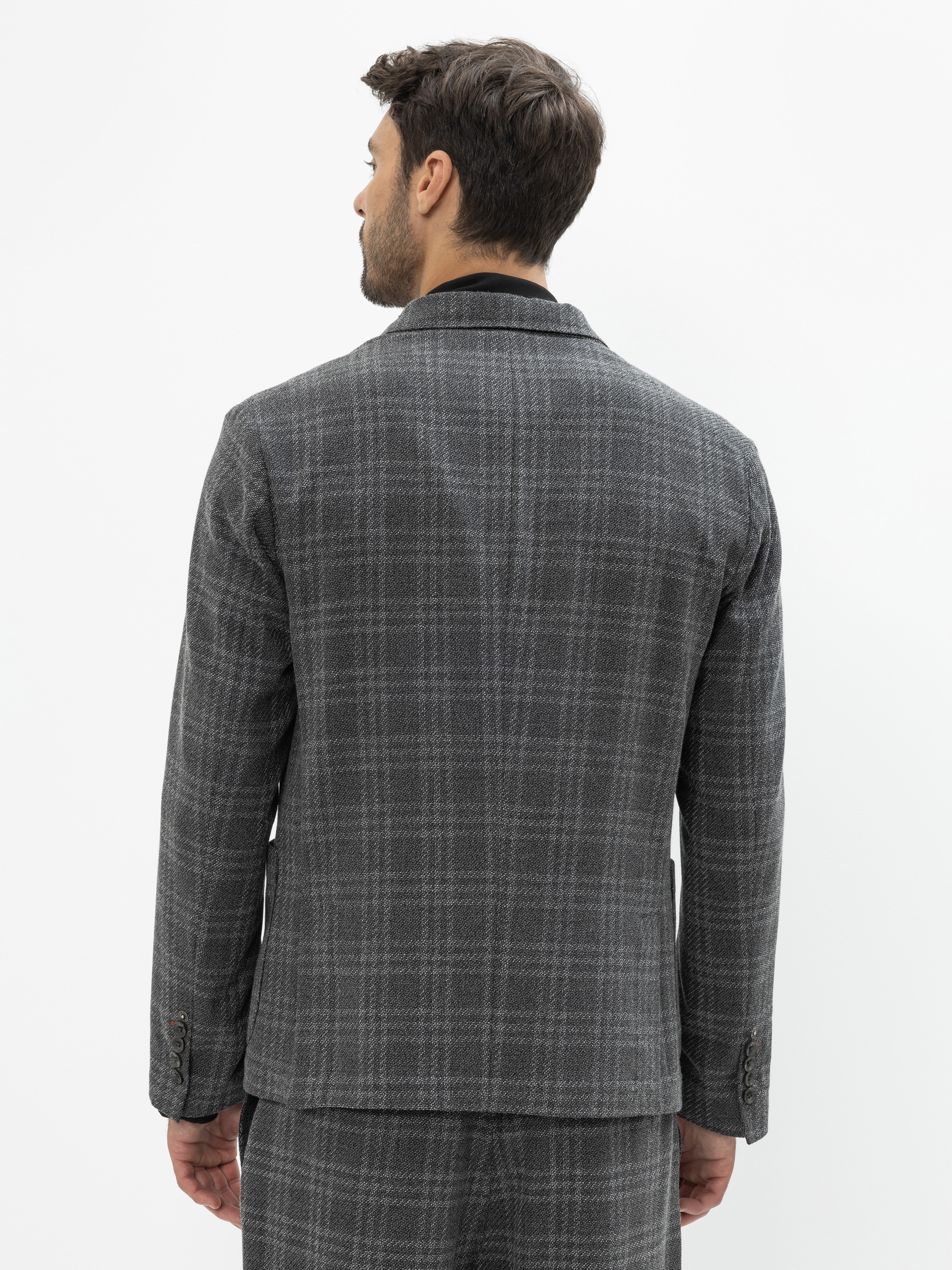 Ein Mann steht mit dem Rücken zur Kamera und trägt eine grau karierte Anzugsjacke und eine dazu passende Hose vor einem schlichten weißen Hintergrund.