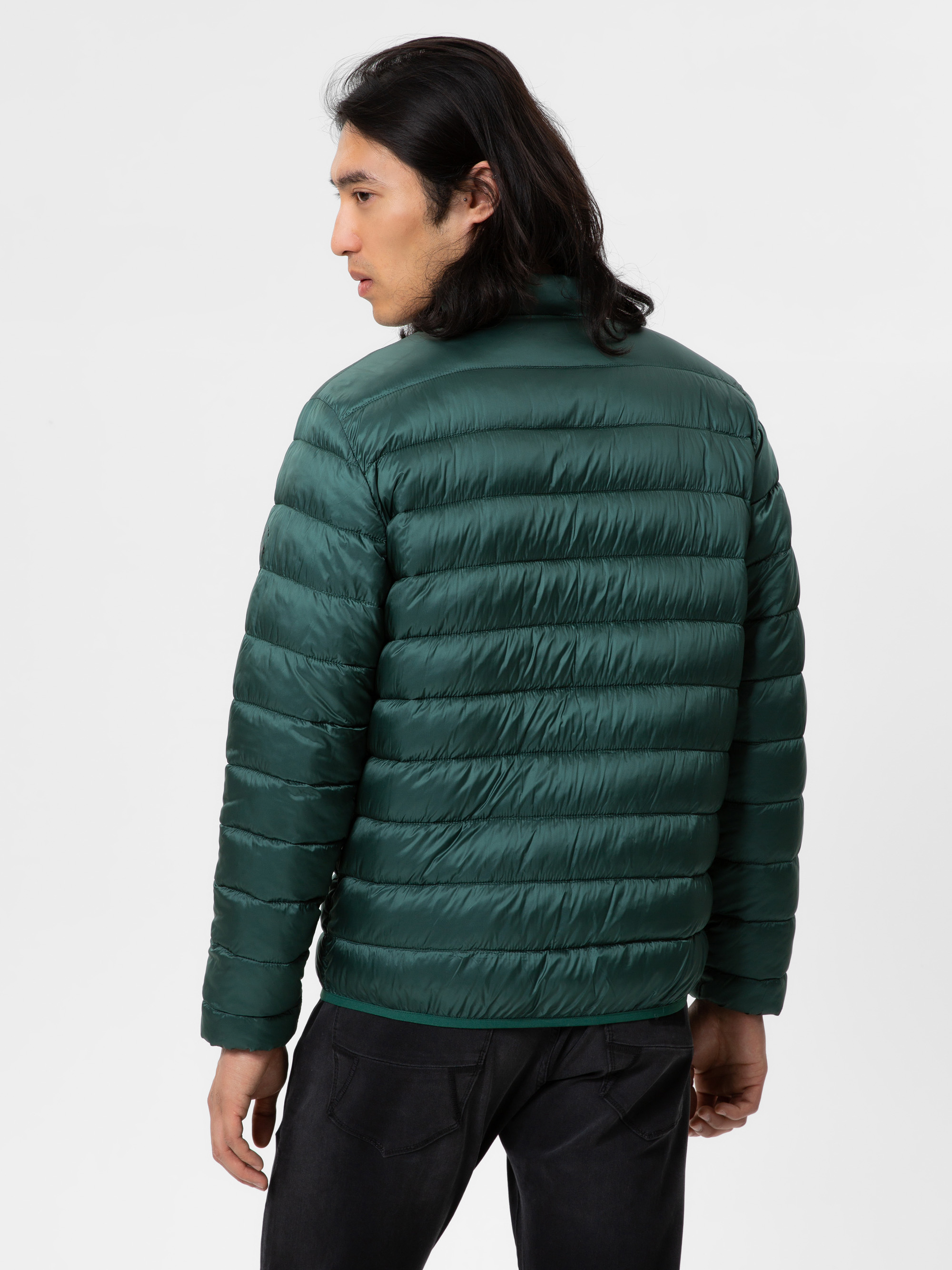 Ein Mann mit langen dunklen Haaren, der eine blaugrüne Steppjacke und schwarze Jeans trägt, steht mit dem Gesicht nach hinten und blickt über seine linke Schulter auf einen schlichten weißen Hintergrund.