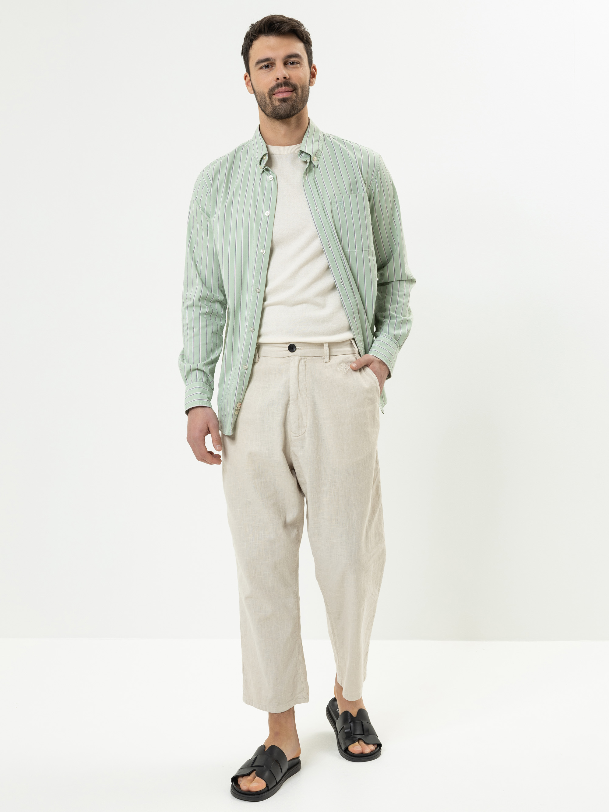 Ein Mann steht vor einem schlichten weißen Hintergrund und trägt ein hellgrün gestreiftes Hemd über einem weißen T-Shirt, eine beigefarbene kurze Hose und schwarze Sandalen. Er hat kurzes dunkles Haar, einen gestutzten Bart und einen entspannten Gesi