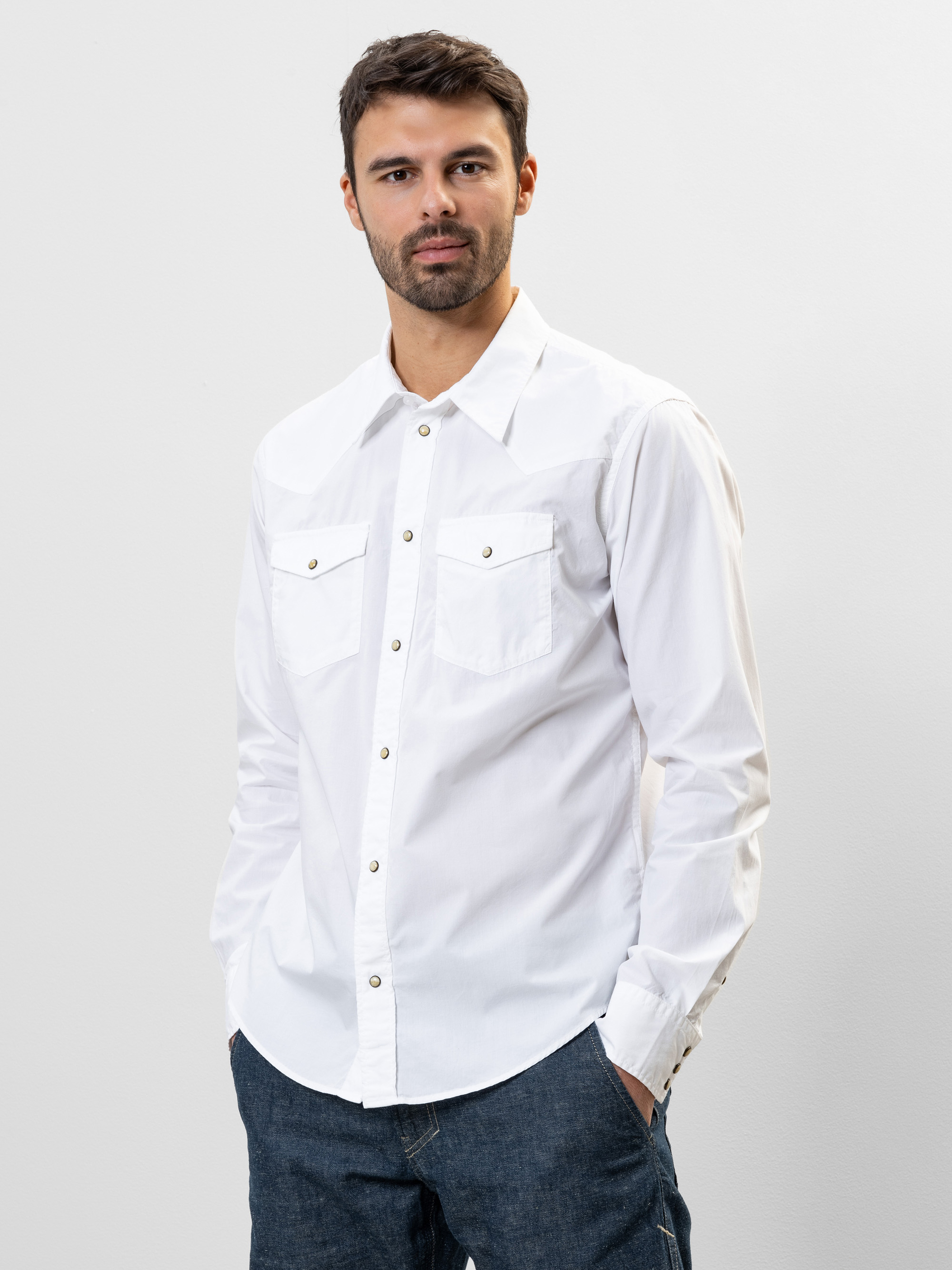 Ein Mann mit kurzen dunklen Haaren und einem Bart trägt ein weißes Button-up-Hemd mit Vordertaschen und blaue Jeans. Er steht vor einem schlichten hellen Hintergrund und hat die Hände in den Taschen.