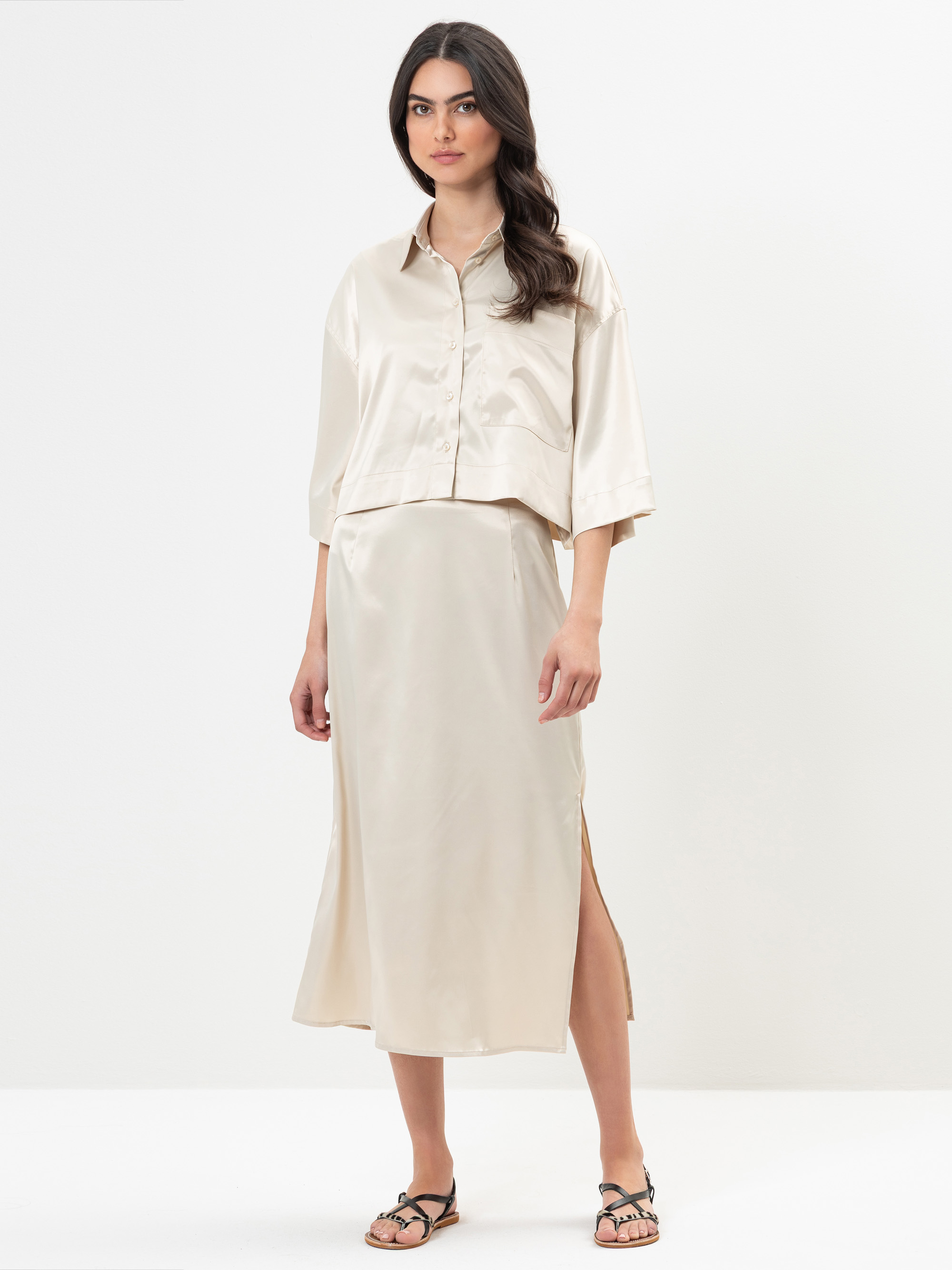 Eine Frau steht vor einem schlichten weißen Hintergrund und trägt ein lockeres, glänzendes, cremefarbenes Button-up-Hemd und einen passenden Midirock mit Seitenschlitz. Sie hat langes dunkles Haar und trägt schwarze Sandalen.