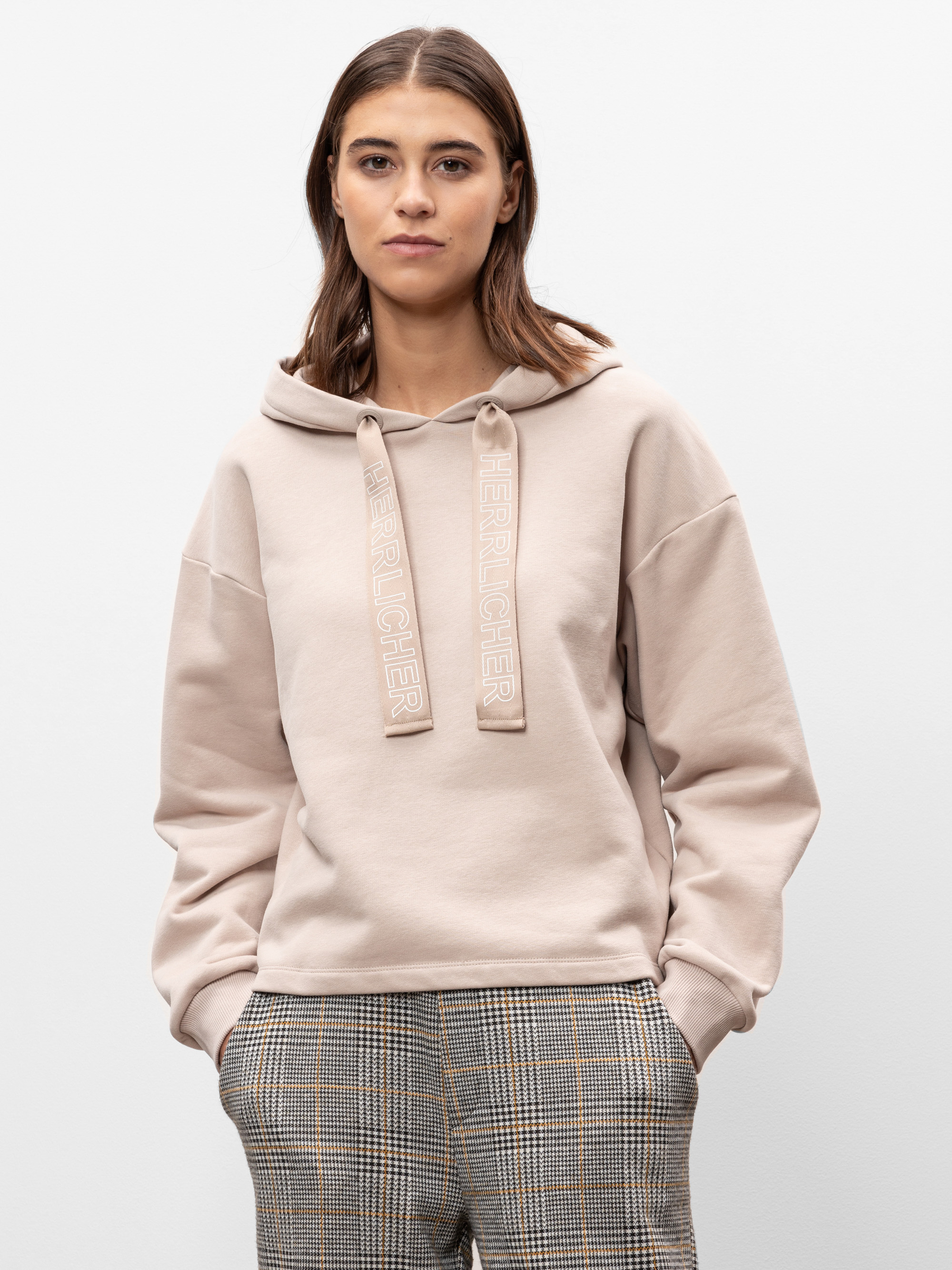 Eine Frau mit glattem braunem Haar trägt einen hellbeigen Kapuzenpulli mit der Aufschrift HER RICHER auf den Kordeln und eine karierte Hose mit hoher Taille. Sie steht mit den Händen in den Taschen vor einem schlichten weißen Hintergrund.