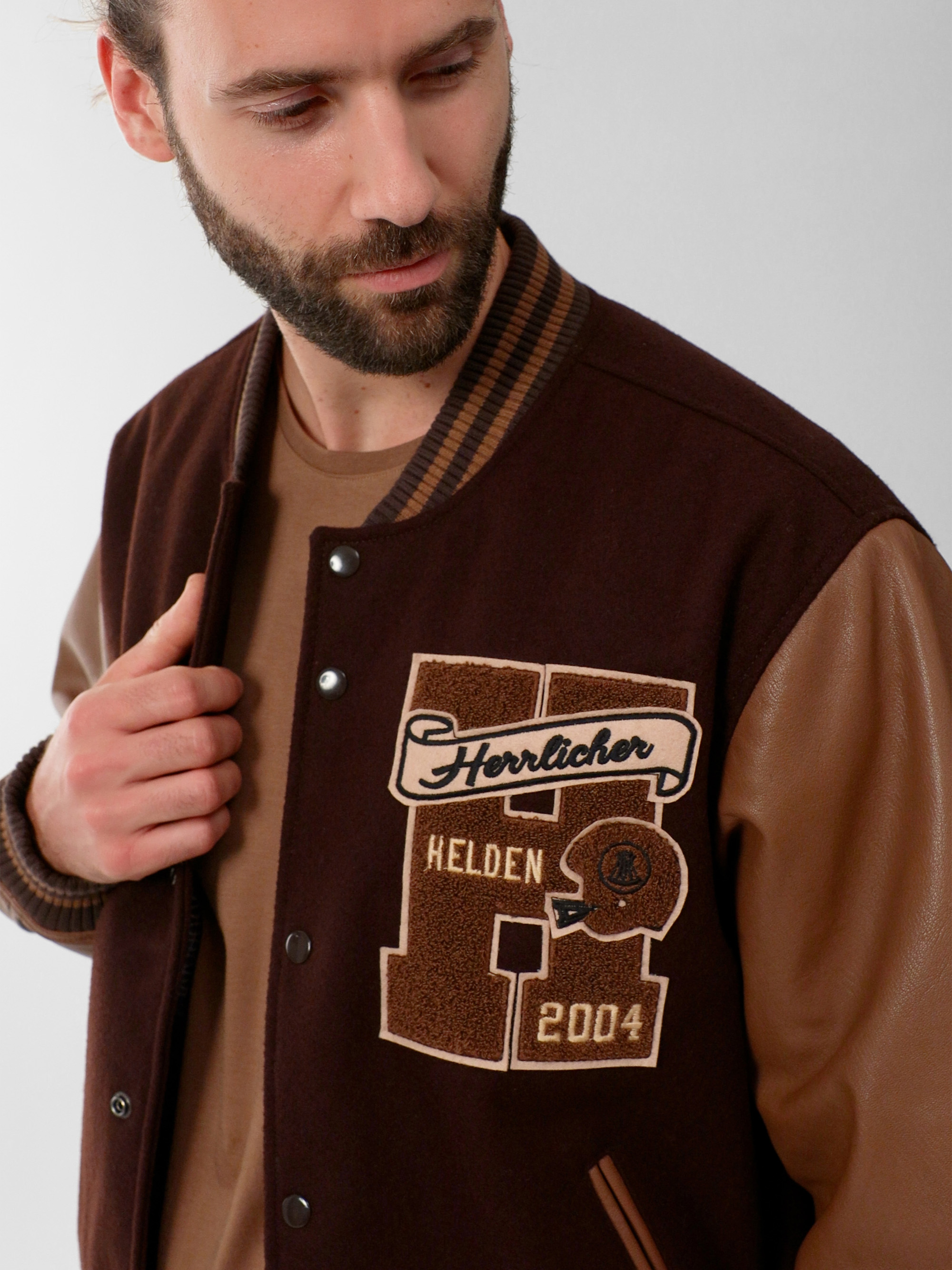 Ein Mann mit Bart trägt eine braune Sportjacke mit hellbraunen Lederärmeln und gestickten Aufnähern mit der Aufschrift "Herrlicher Helden 2004". Er blickt nach unten und leicht zur Seite.