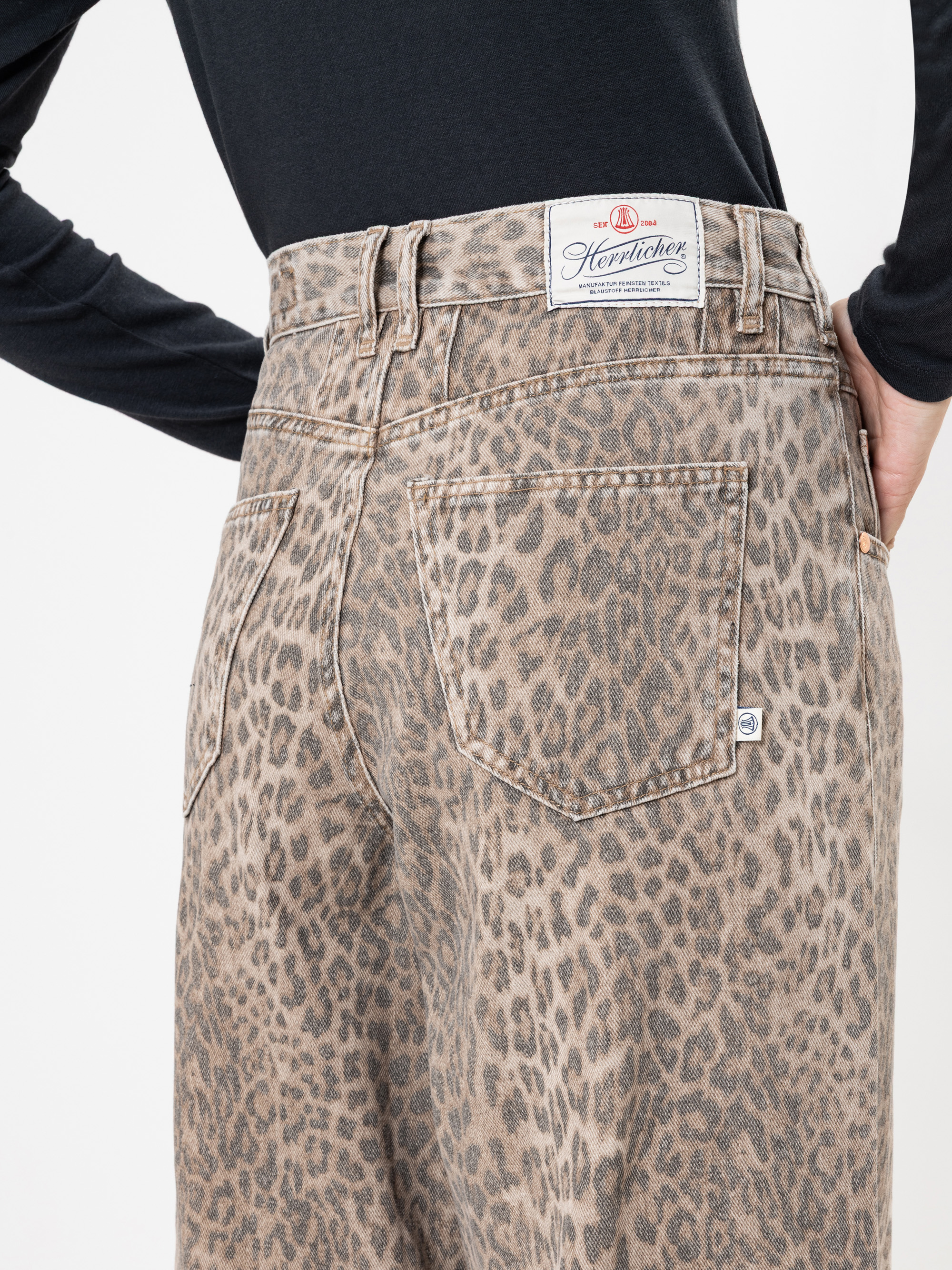 Person, die eine Jeans mit hoher Taille und Leopardenmuster trägt, mit einem weißen Herrlicher-Markenetikett am Bund. Die Person ist mit einem schwarzen langärmeligen Oberteil bekleidet, das von hinten und leicht zur Seite gedreht gezeigt wird.