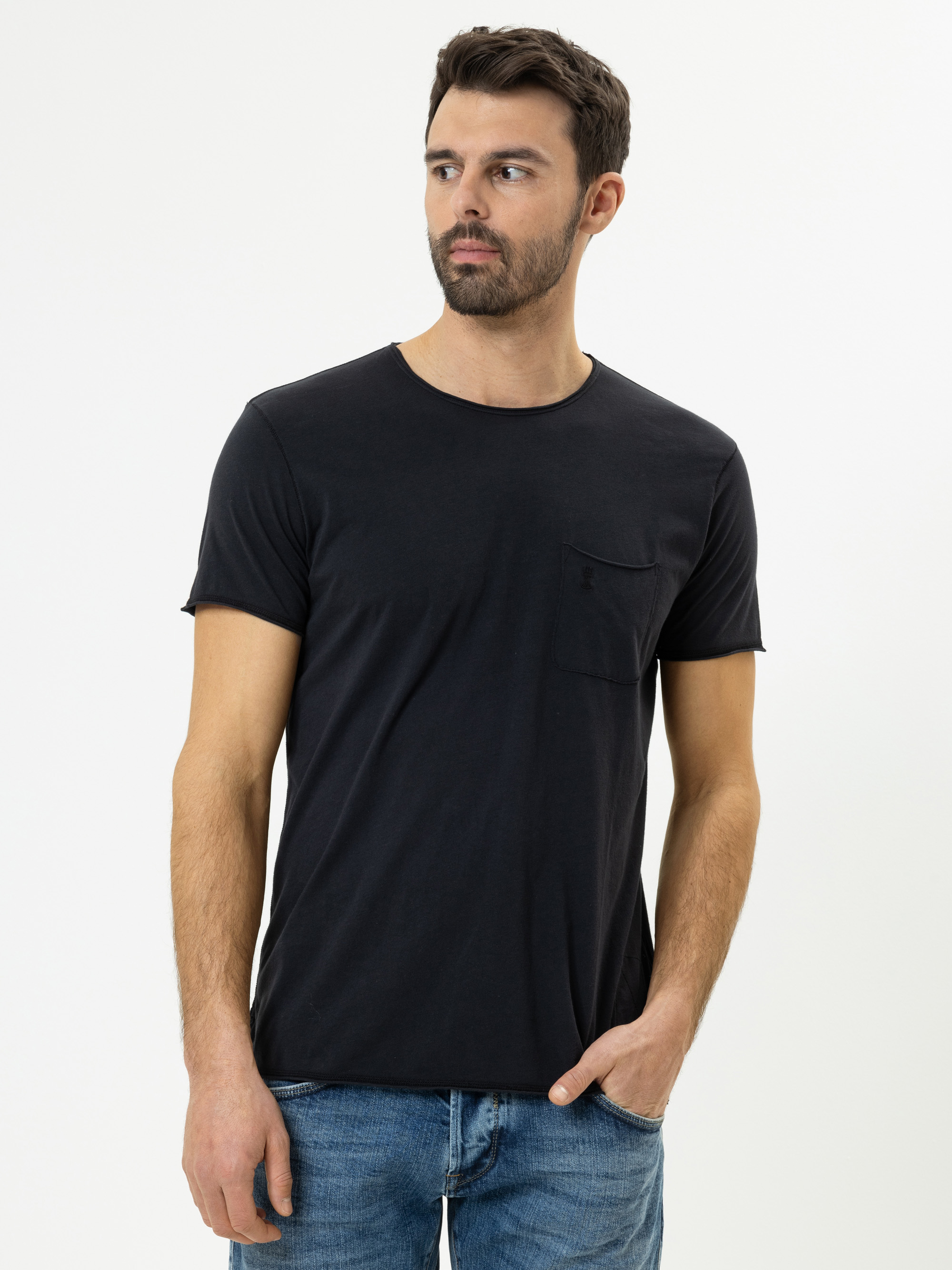 Ein Mann mit kurzen dunklen Haaren und Bart trägt ein schlichtes schwarzes T-Shirt mit einer Brusttasche und blaue Jeans. Er steht vor einem weißen Hintergrund, blickt leicht nach links und hat eine Hand in der Tasche.