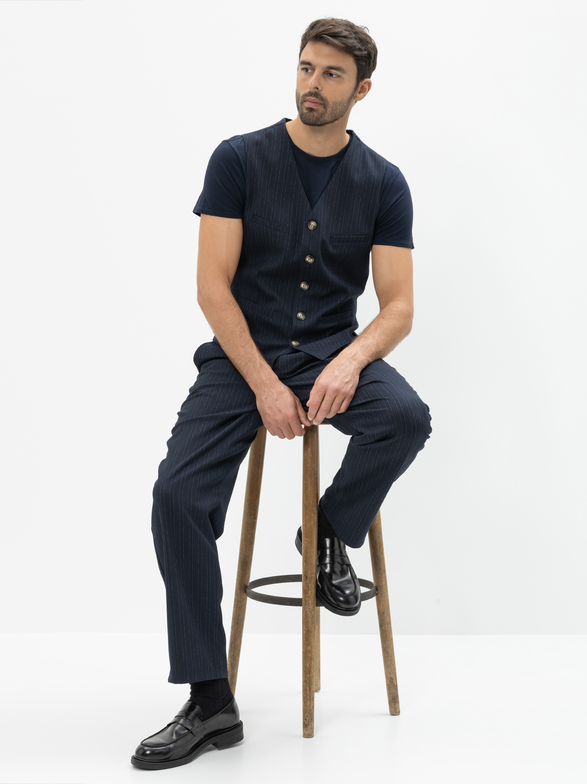 Ein Mann mit kurzen dunklen Haaren und einem Bart sitzt auf einem Holzschemel vor einem weißen Hintergrund. Er trägt eine marineblaue Nadelstreifenweste und eine passende Hose, ein dunkles T-Shirt und schwarze Anzugschuhe.