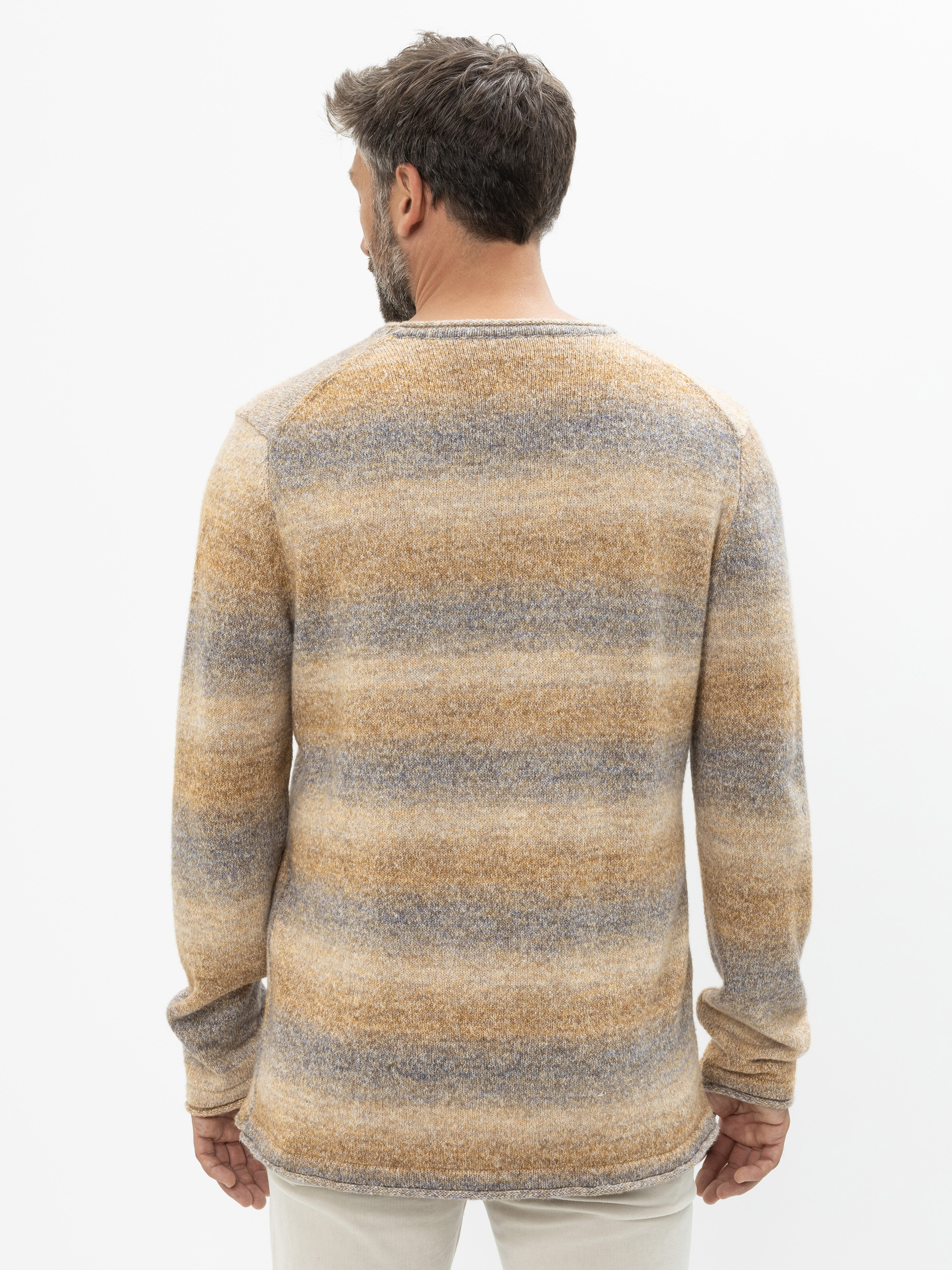 Ein Mann mit kurzen braunen Haaren und einem Bart ist von hinten zu sehen. Er trägt einen langärmeligen Pullover mit horizontalen Streifen in Beige-, Braun- und Grautönen und eine helle Hose vor einem einfarbig weißen Hintergrund.
